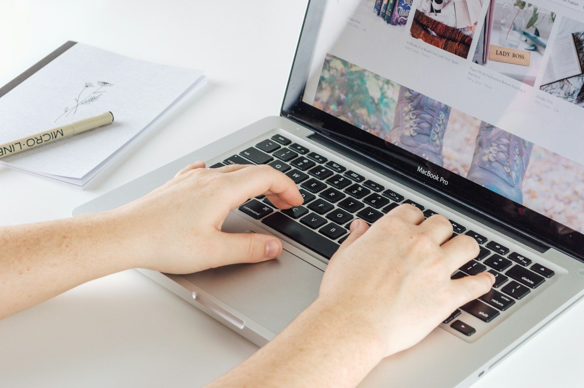 Hands typing on a silver laptop on a white desk with a notepad and pen.