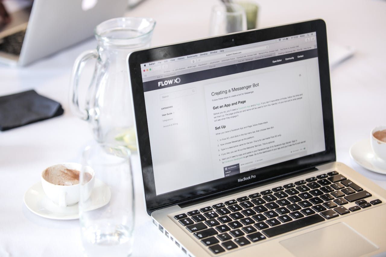 Laptop displaying a webpage, on a white tablecloth, with a pitcher of water and a cup of coffee nearby.