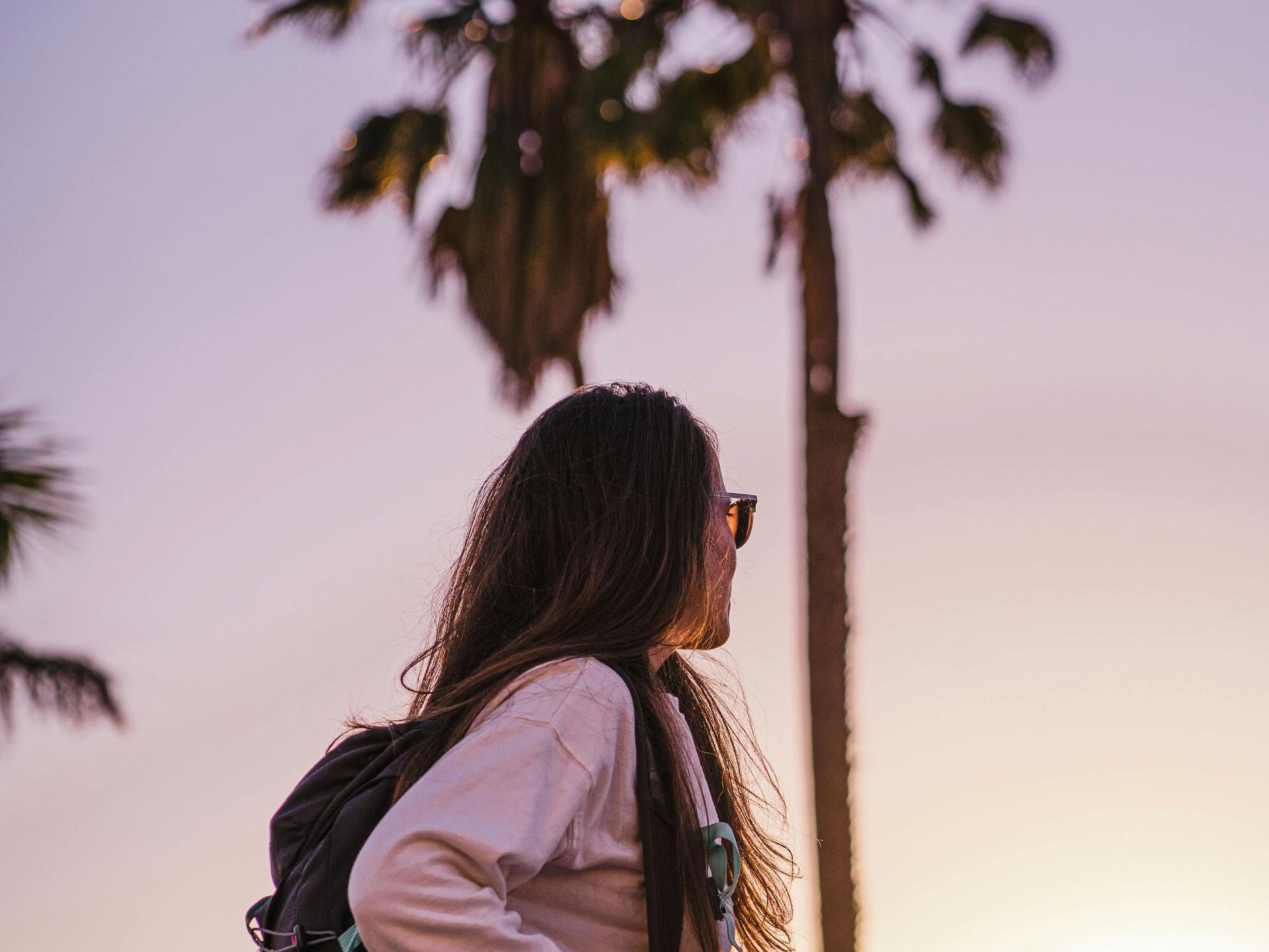 Woman with backpack under palm trees at sunset.
