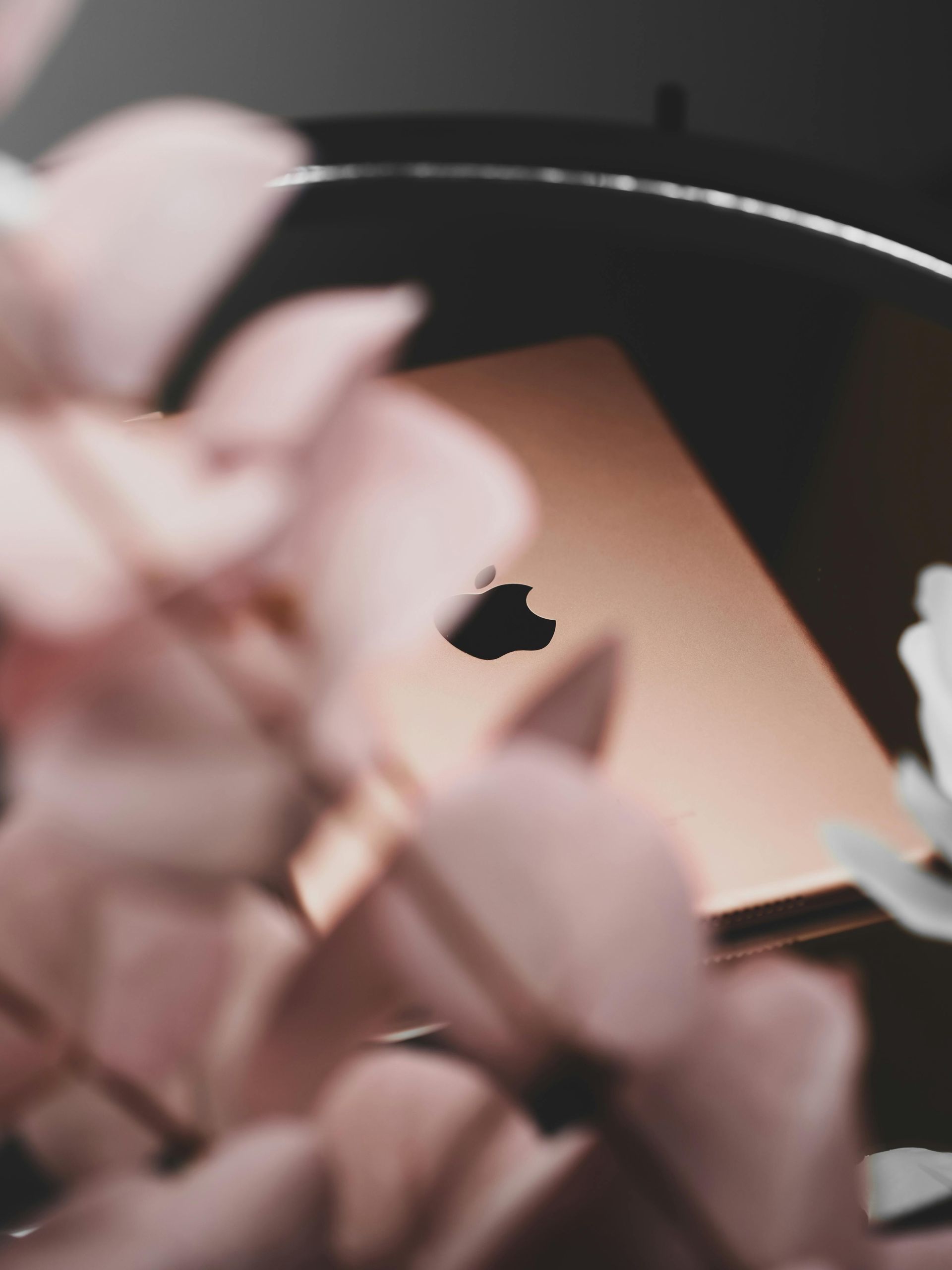 Rose-gold laptop with black Apple logo, set against a dark background and partly obscured by pink flowers.