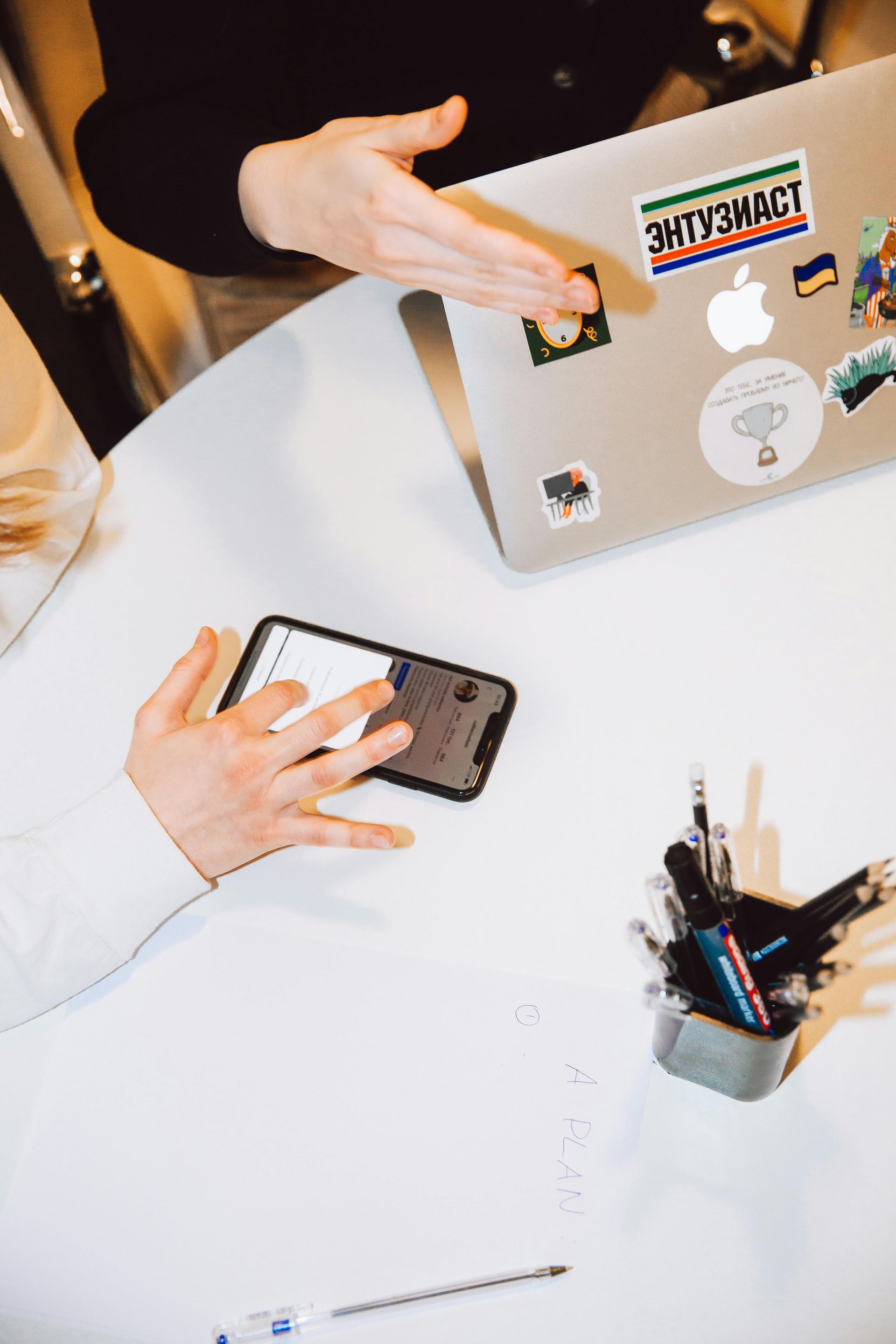 Hands interacting with a smartphone and pointing at a laptop on a white table. A pen holder and tools are also present.
