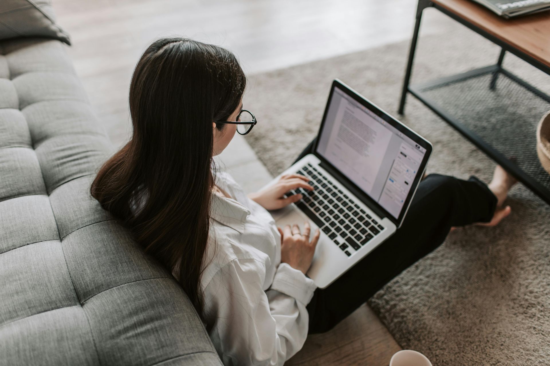 Woman with glasses typing on a laptop, seated on the floor near a couch.