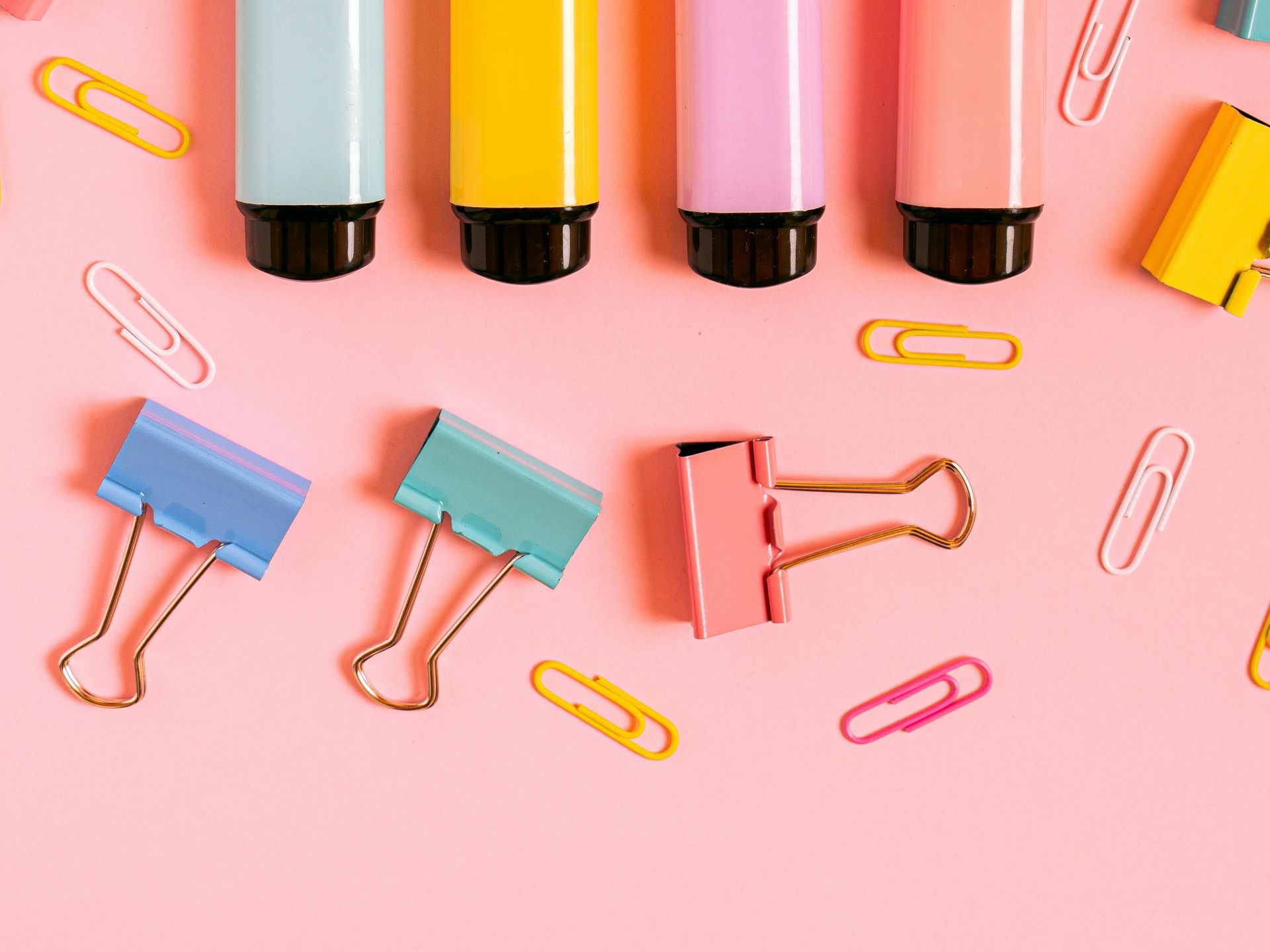 Colorful highlighters, binder clips, and paper clips on a pink background.