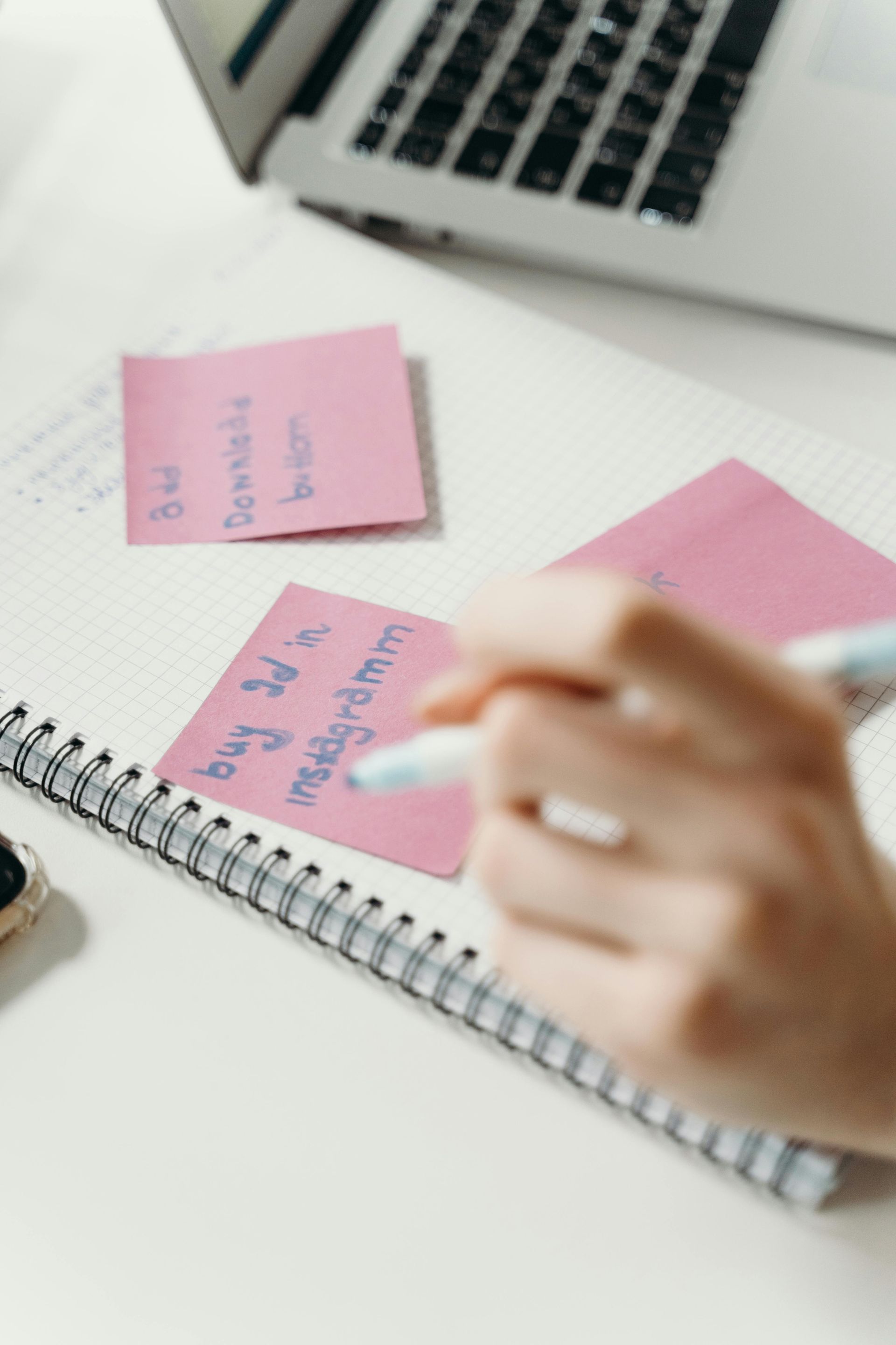 Person writing notes on pink sticky notes, notebook and laptop on a white surface.