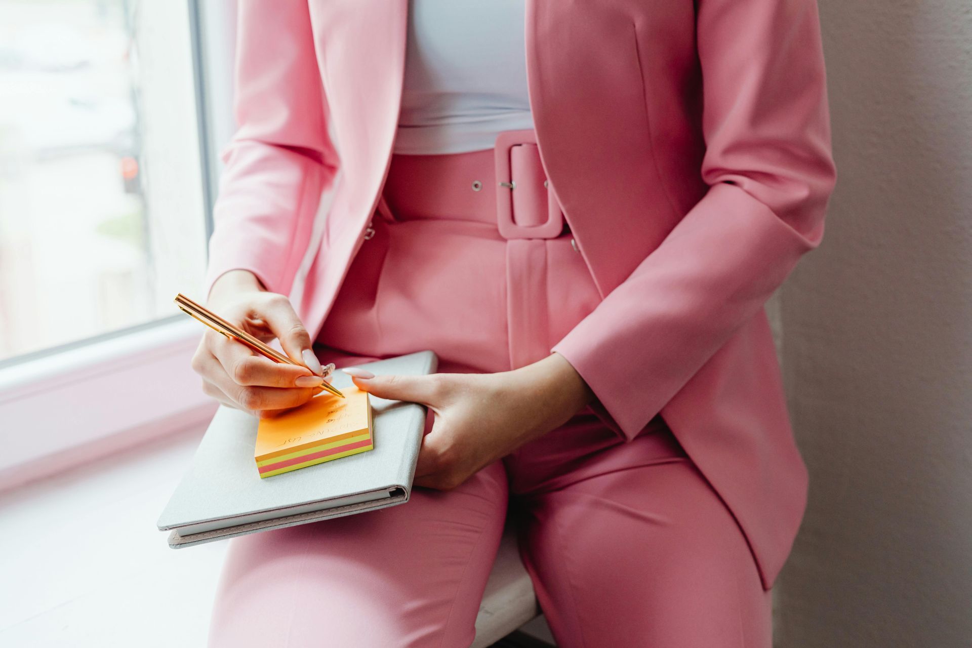 Person in pink suit writing in a notebook with sticky notes by a window.