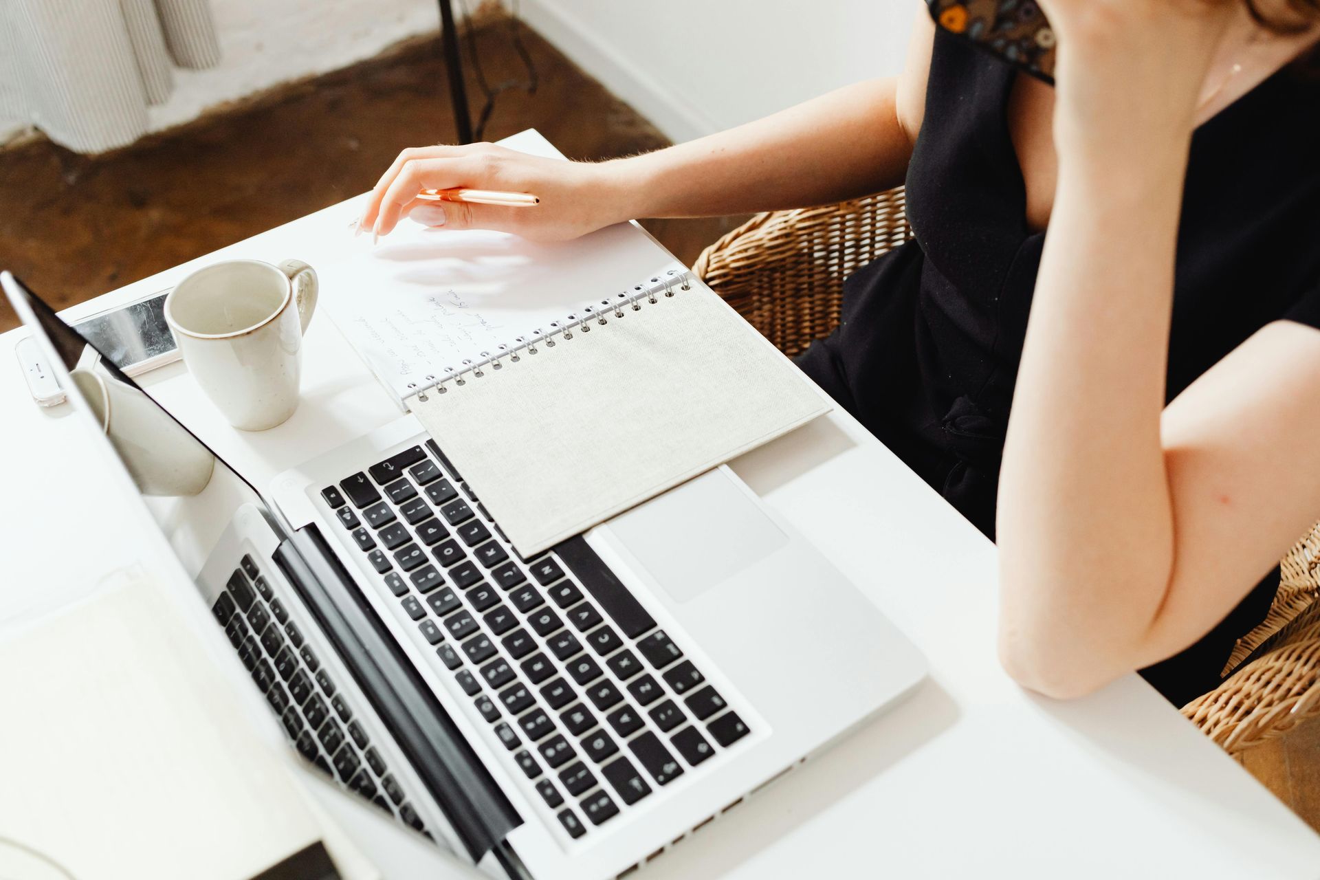 Woman at desk with laptop, notebook, mug. She rests chin on hand. Bright room.