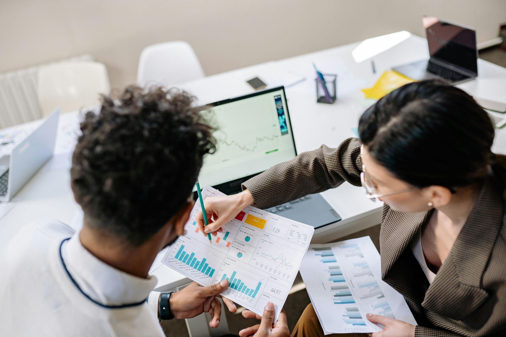 Two people reviewing charts on a laptop and paper at a white office desk. Woman points to a chart.