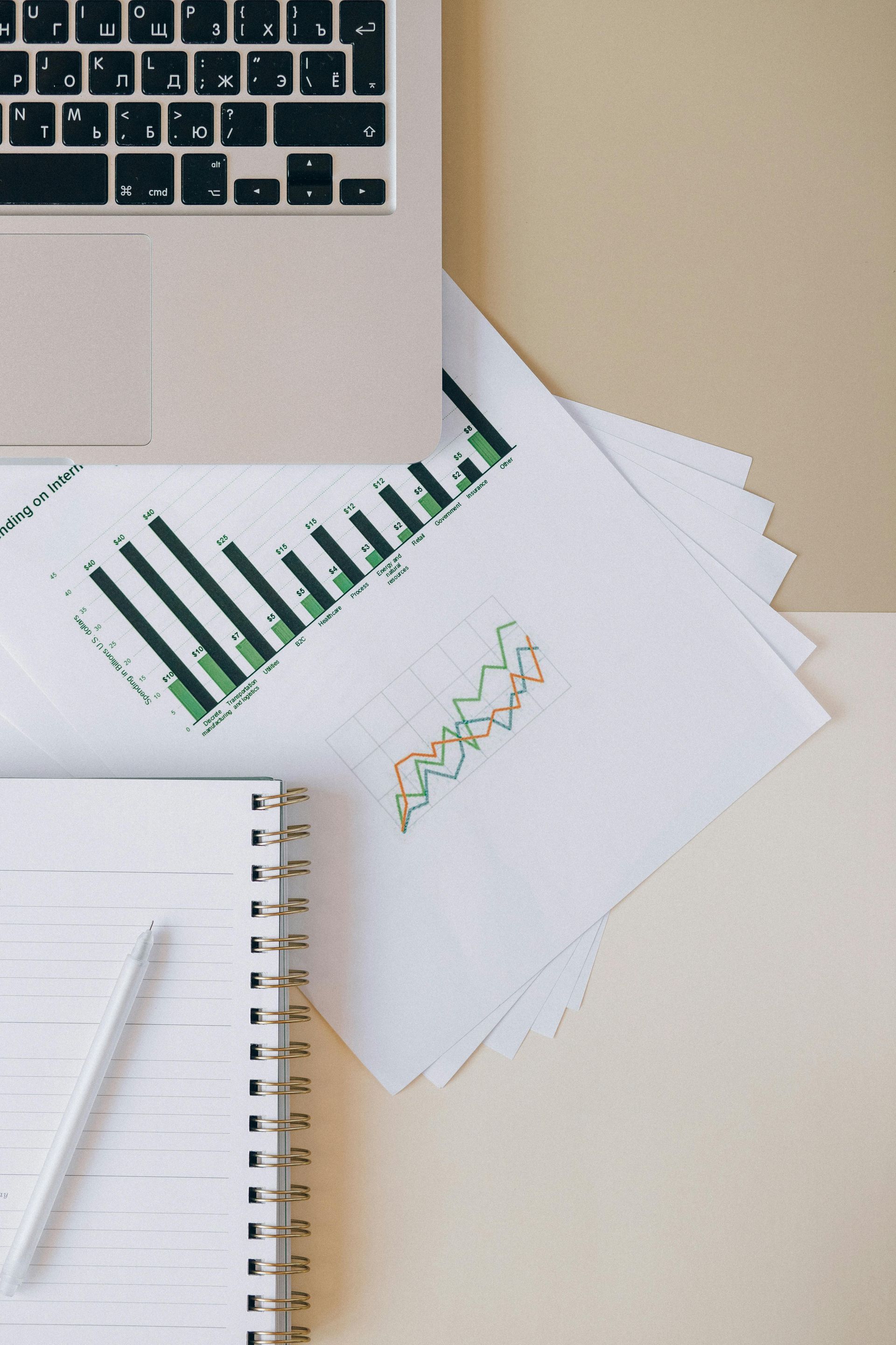 Laptop, charts, notepad, and pencil on a light beige surface.