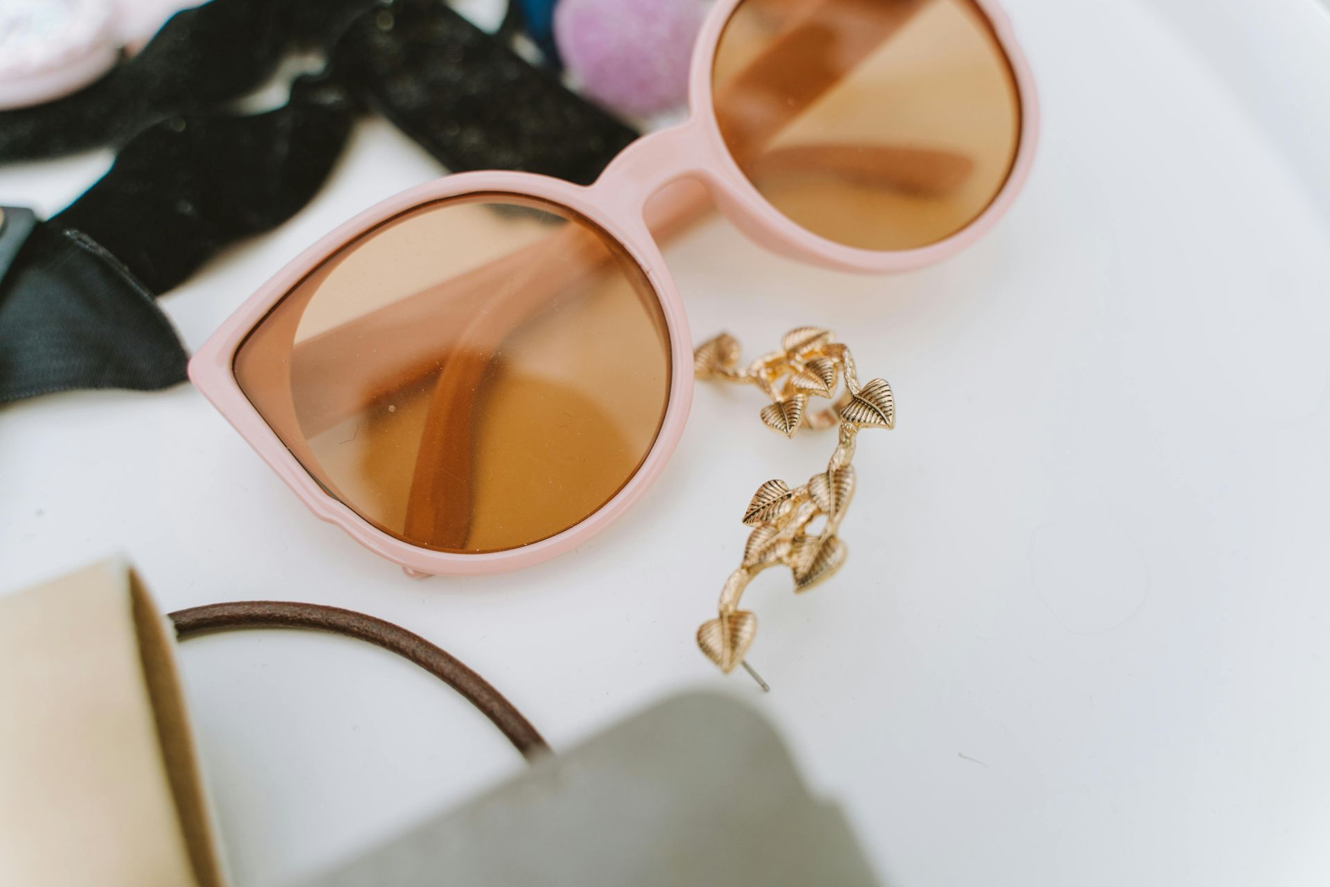 Pink sunglasses, gold earrings, hair ties, and a small box on a white surface.