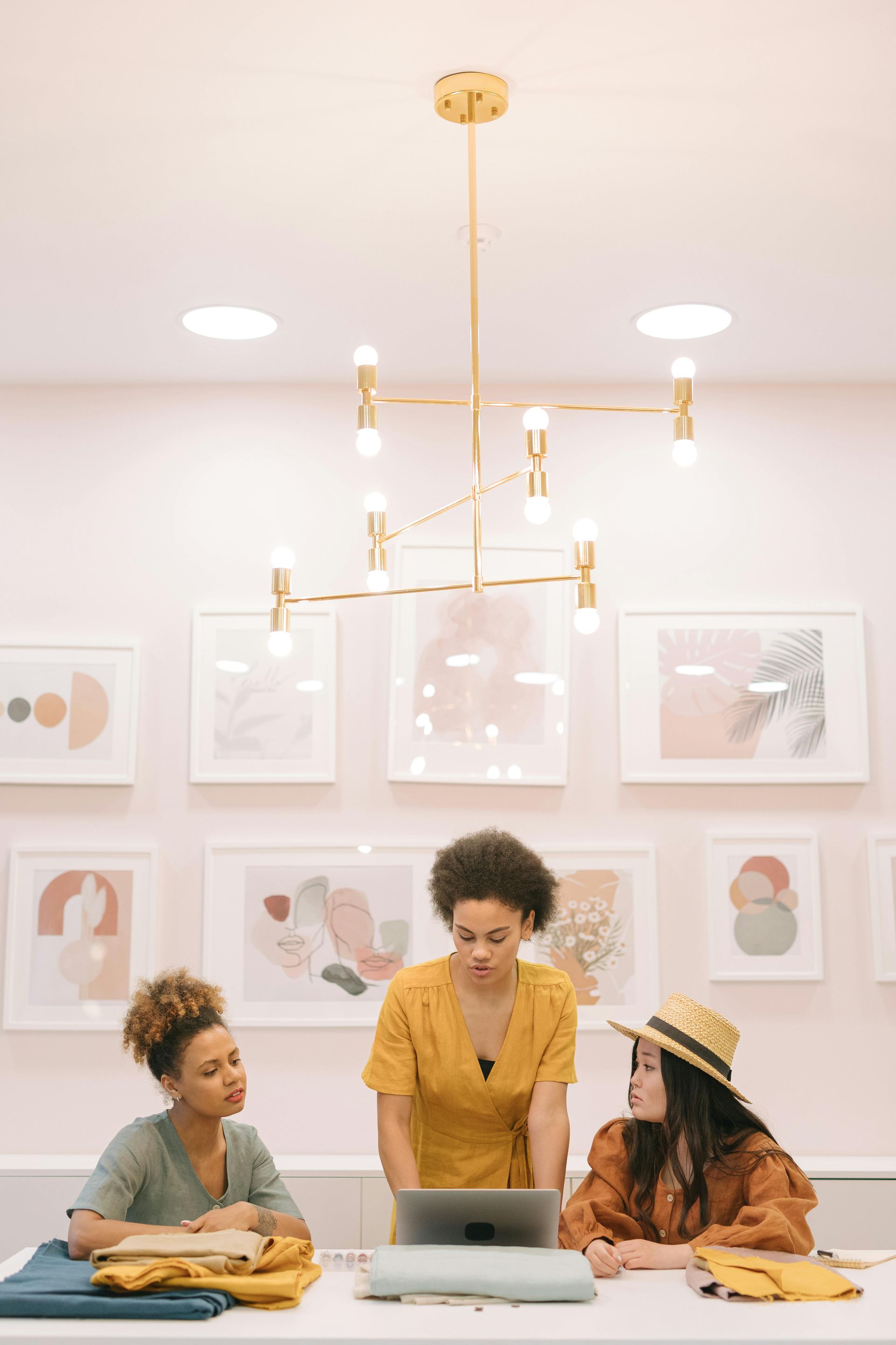 Three women at a table looking at a tablet, light pink room, framed art on the wall, gold chandelier.