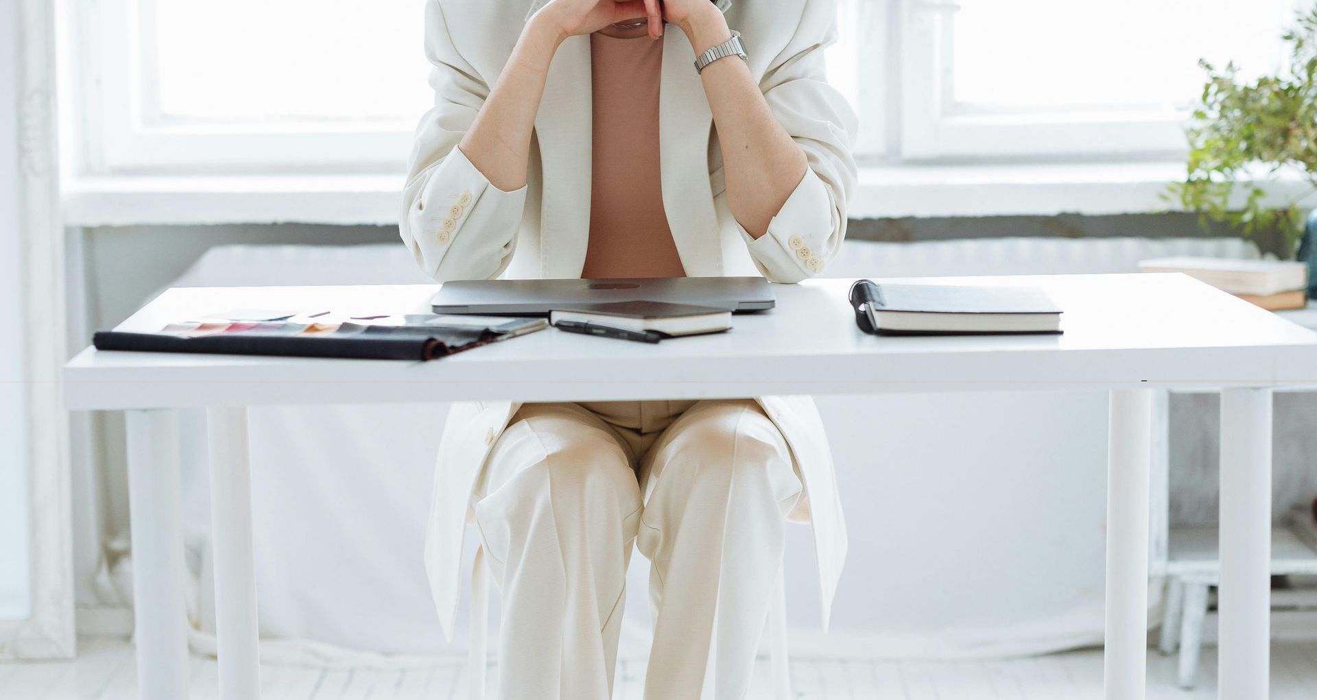Woman in white suit at a desk, hands clasped near face, laptop and notebook present.