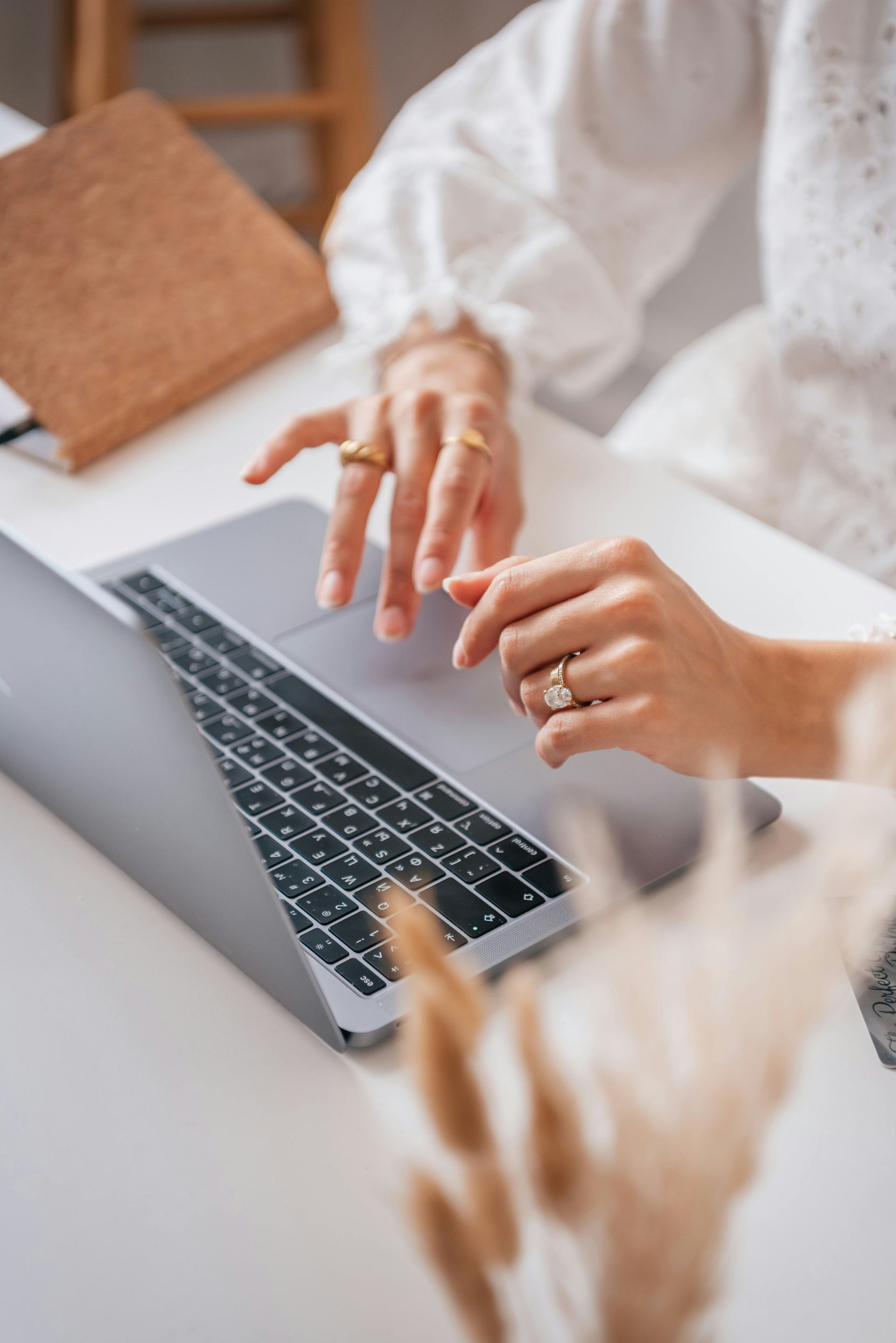 Woman's hands on laptop keyboard, typing, wearing rings, white top, neutral setting.