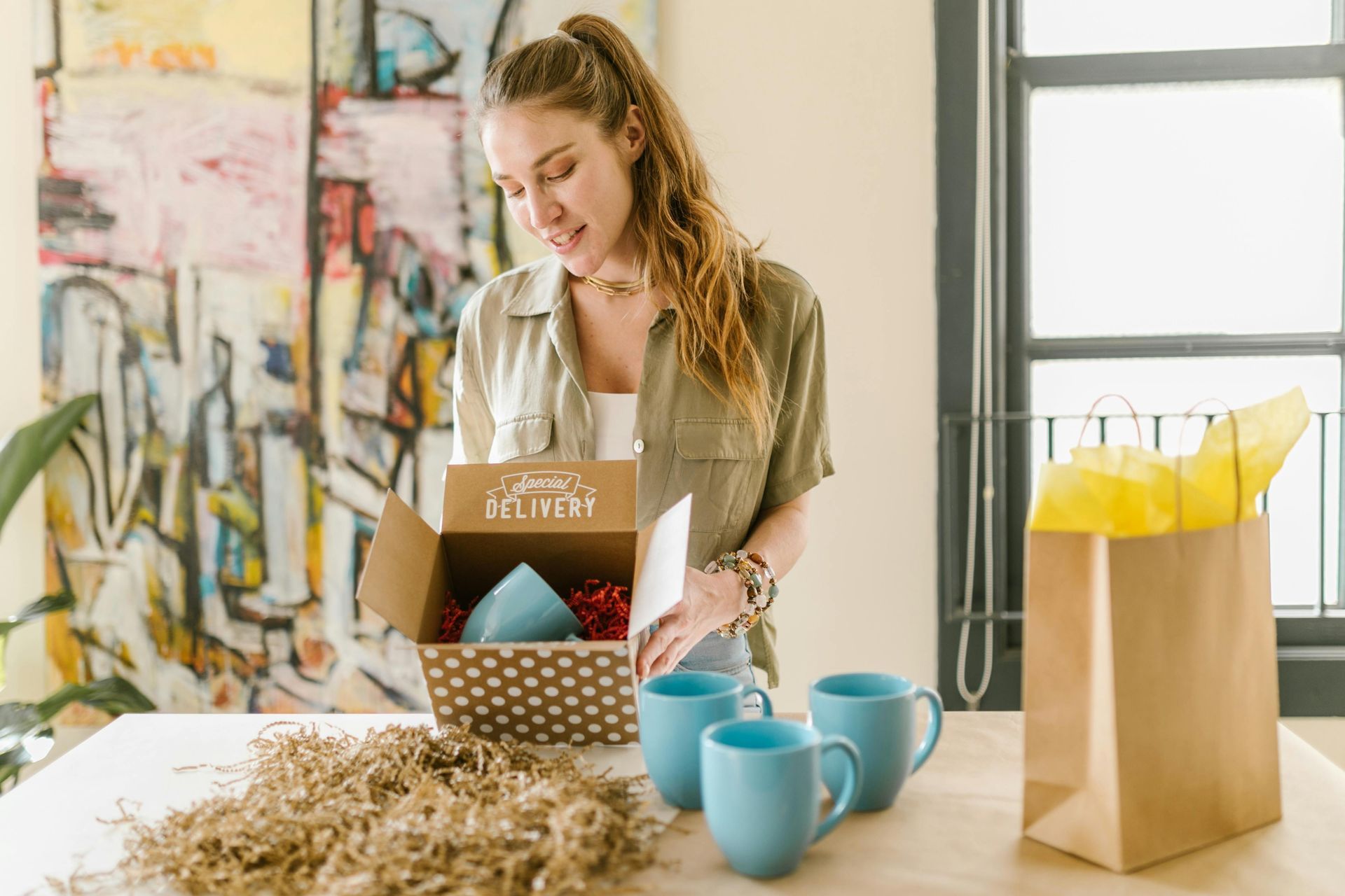 Woman opening a gift box with blue mugs, table with shredded paper, gift bag, abstract art in the background.