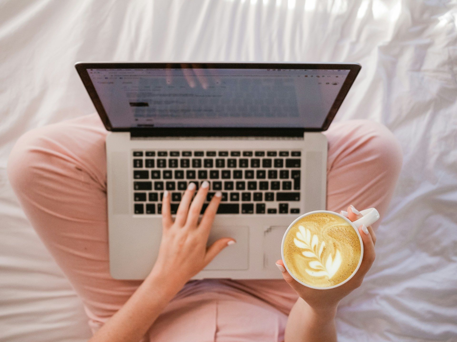 Person in pink pants using a laptop on a bed, holding a latte with a leaf design.