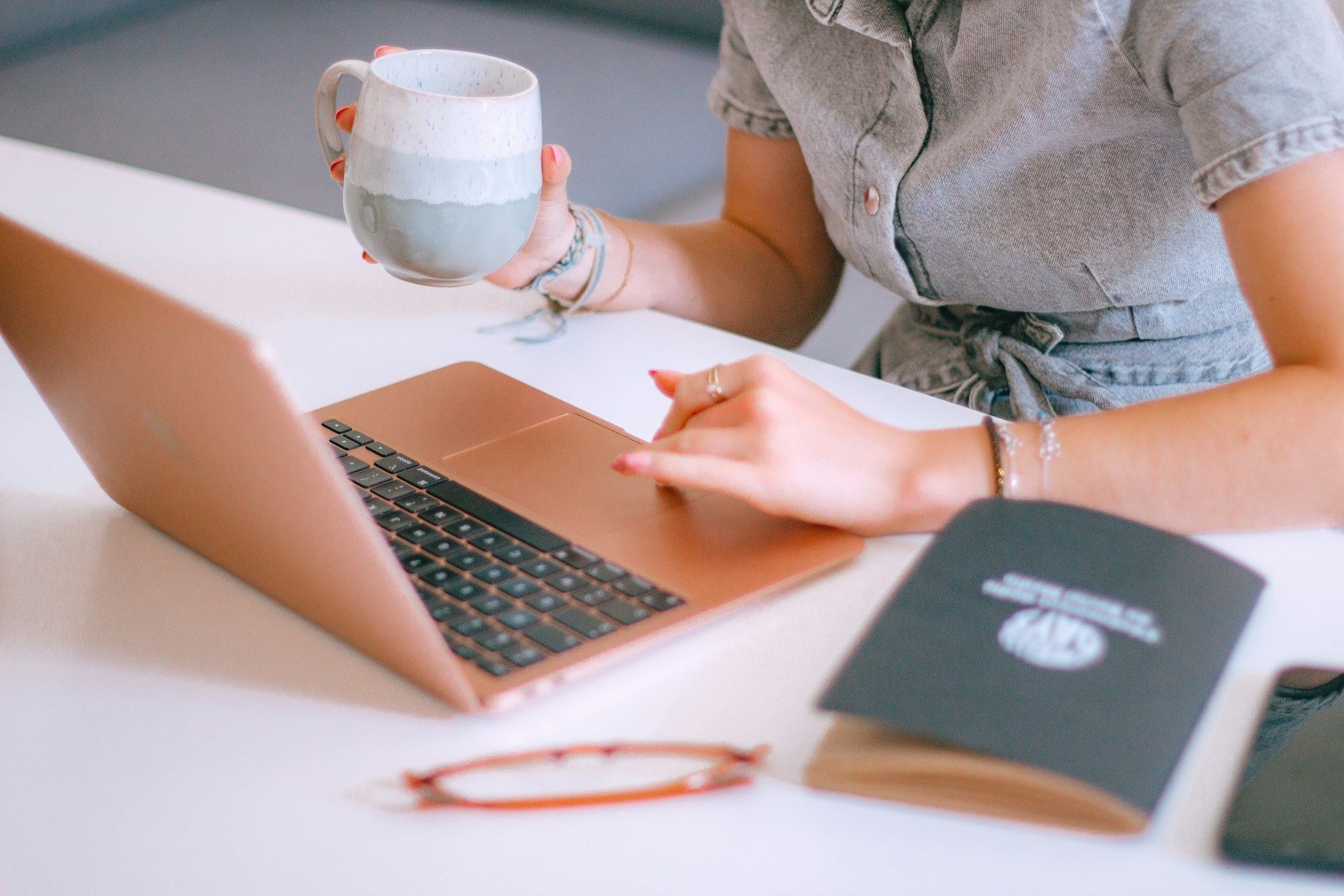 Woman at desk with laptop, holding mug, touching laptop touchpad. Notebook and phone on desk.