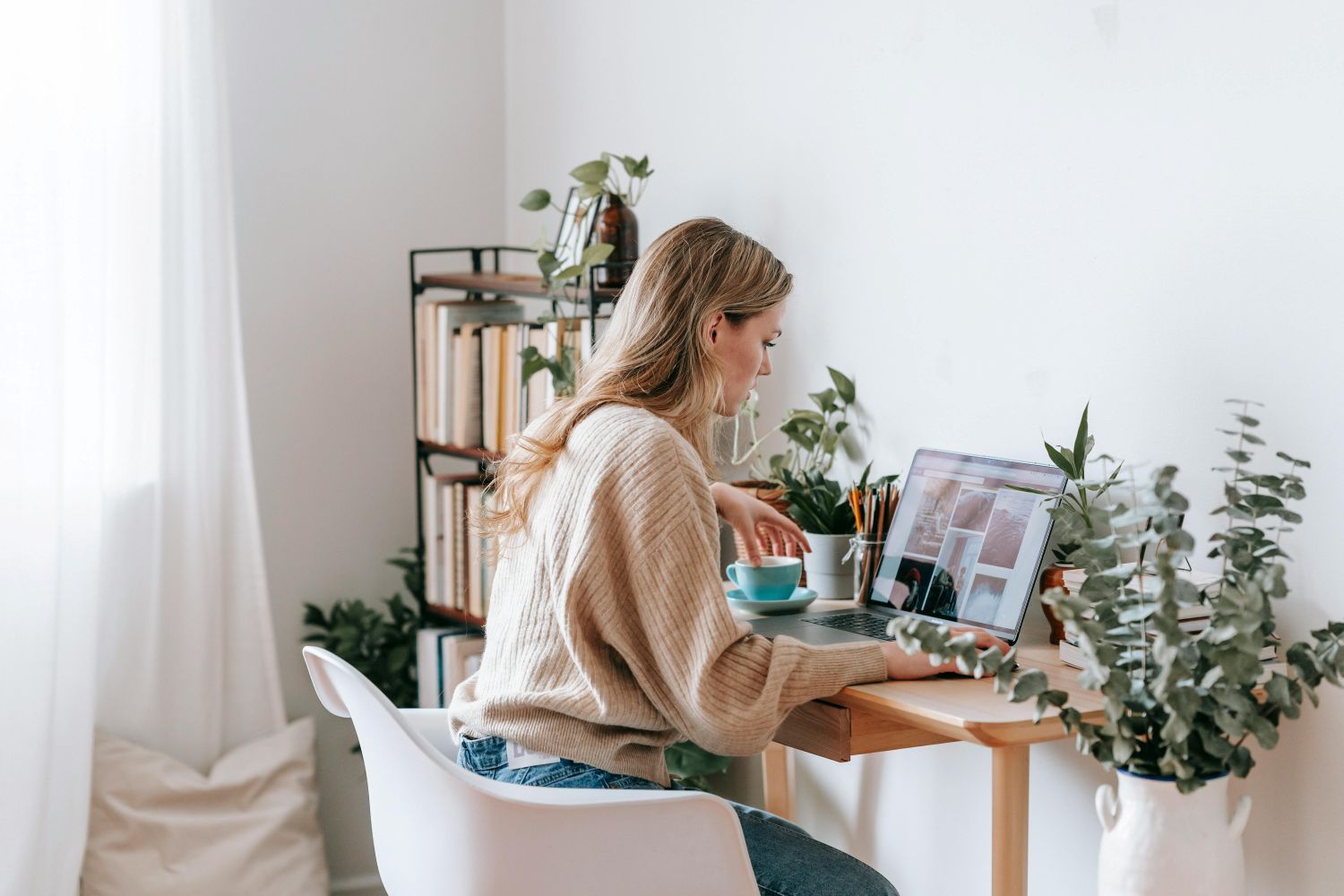 Woman in sweater working on laptop at a desk with plants and a cup, in a bright room.
