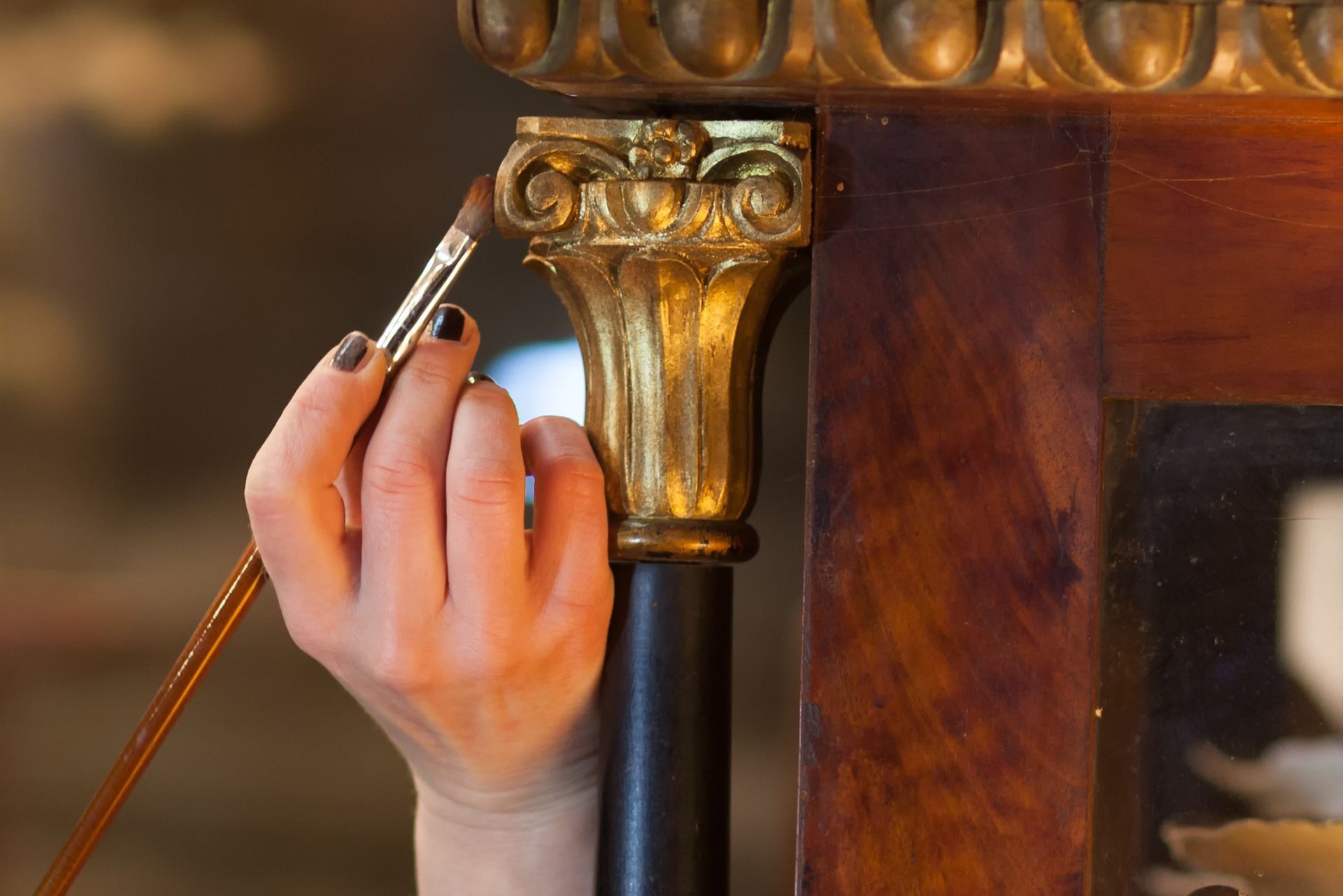A close-up of a person using a thin brush to carefully paint or gild a small, ornate golden column detail on furniture.