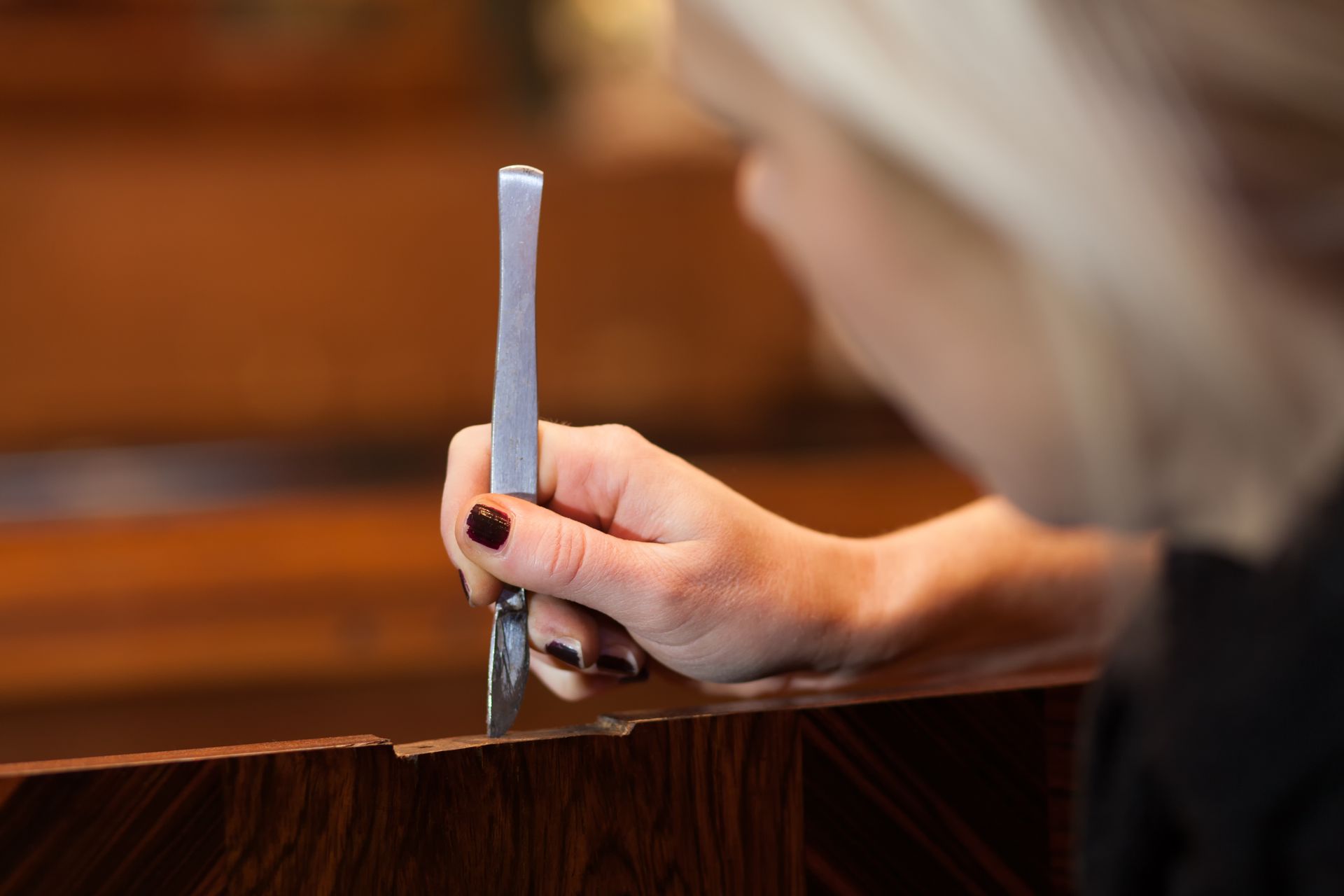 A person uses a scalpel to carefully trim or repair the edge of a piece of dark, wood-grained furniture.