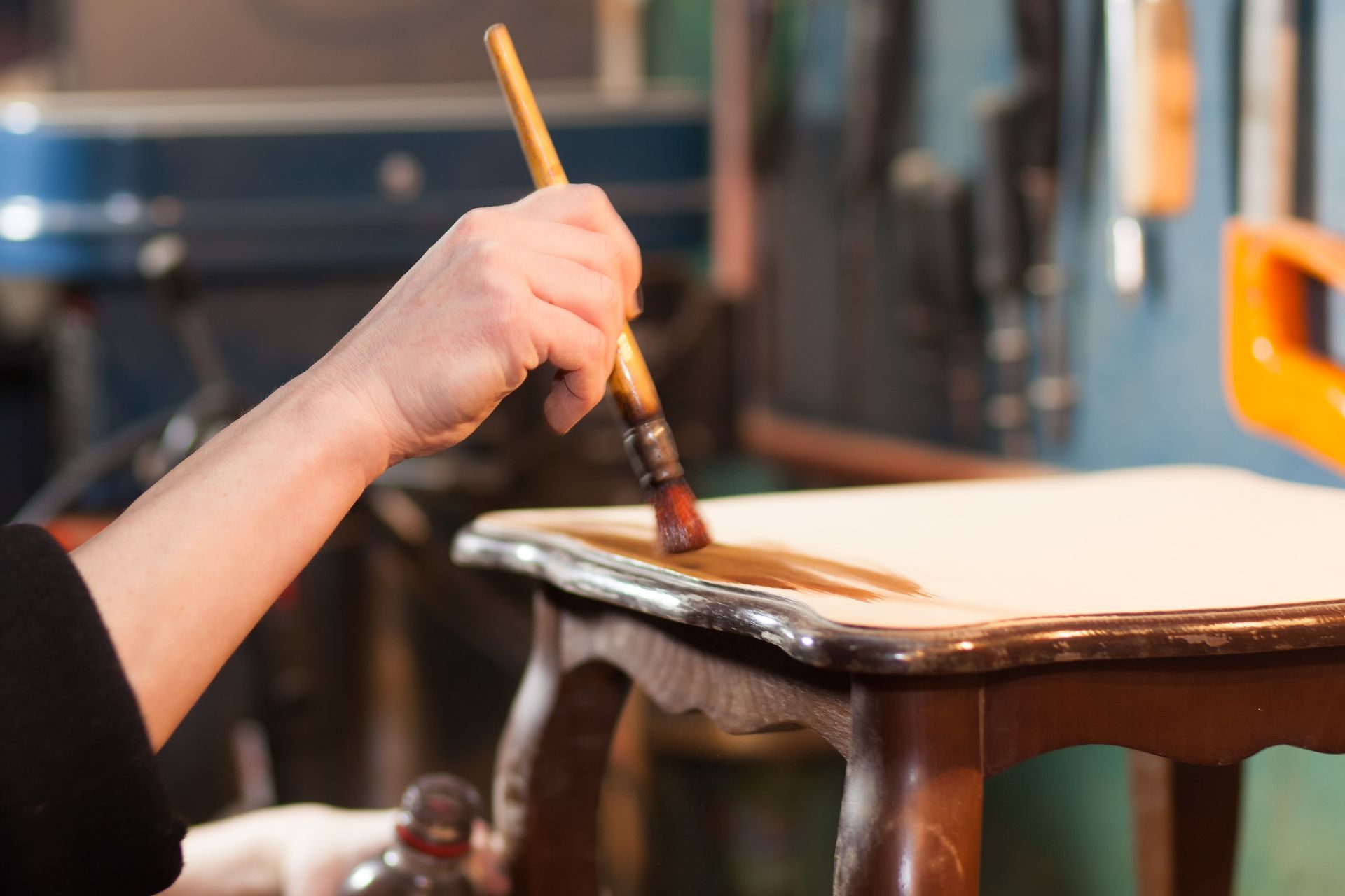 A person applies wood stain to the top of a small, wooden table with a paintbrush in a workshop.