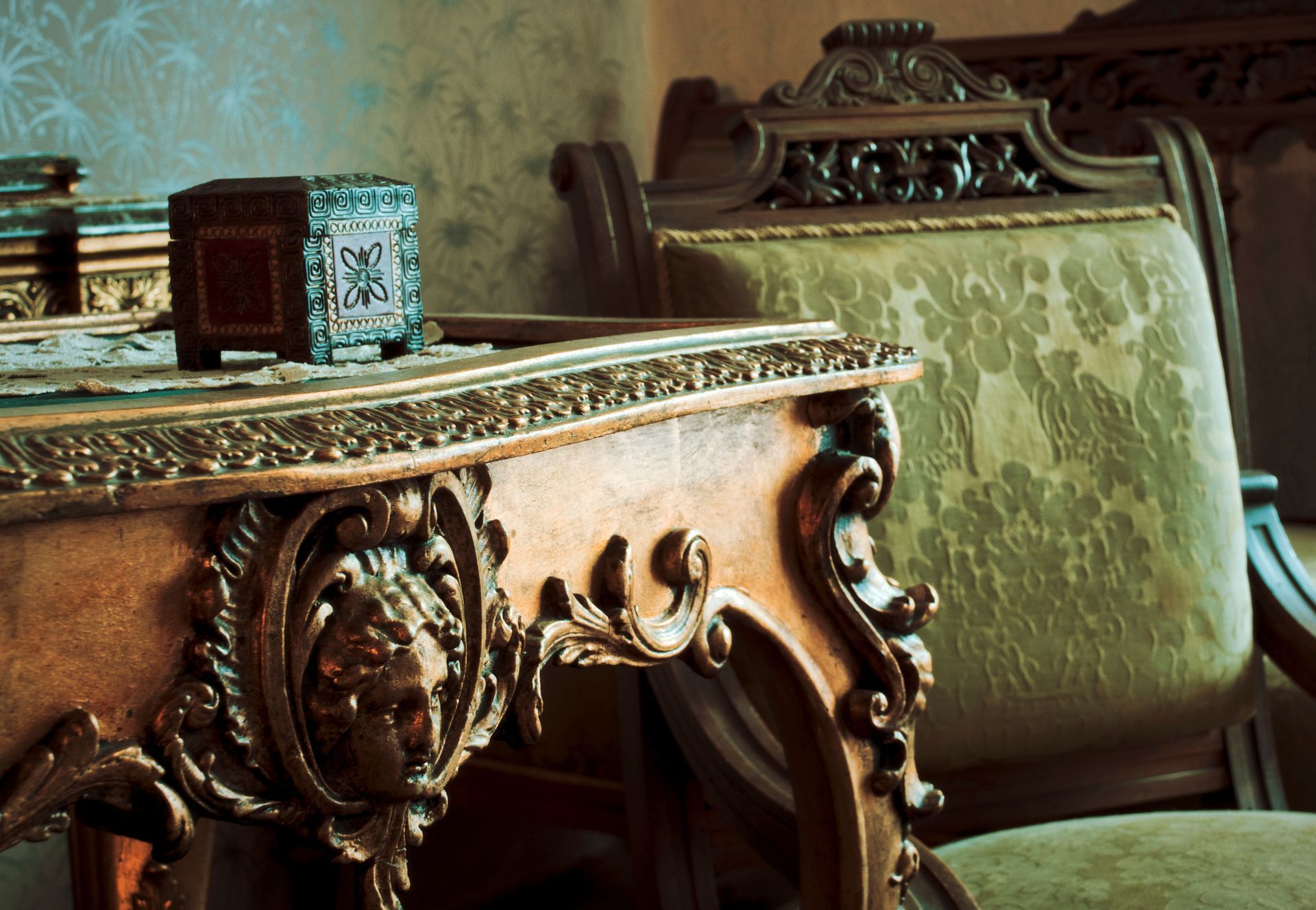 A carved wooden table with an ornate face design sits next to a vintage, upholstered green chair.