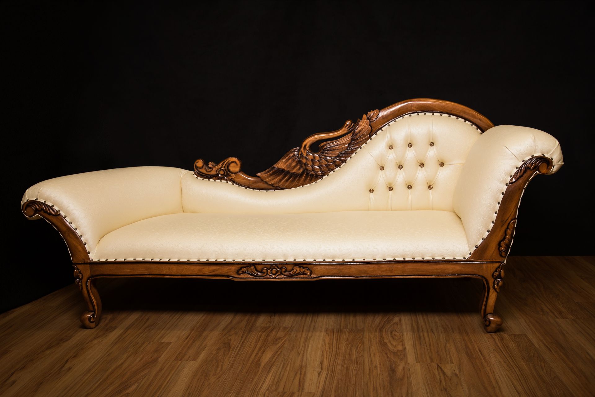 An elegant, cream-colored tufted fainting couch with ornate, dark wood carvings, set against a dark background.