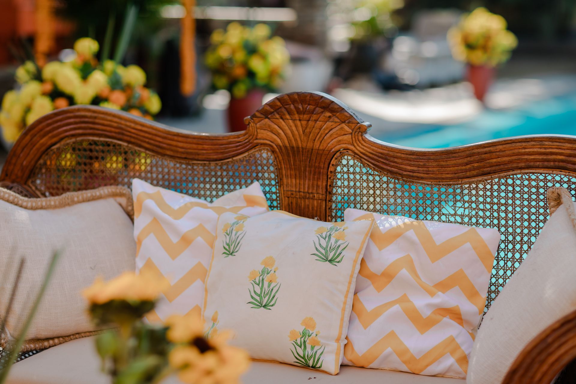A vintage wooden cane sofa outdoors, decorated with white and yellow chevron and floral print cushions near a pool.