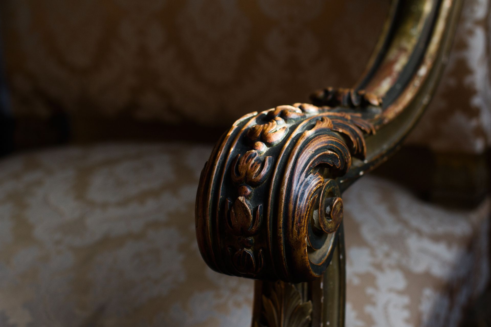 Close-up of a carved wooden armrest with gold detailing on an upholstered vintage chair with patterned fabric.