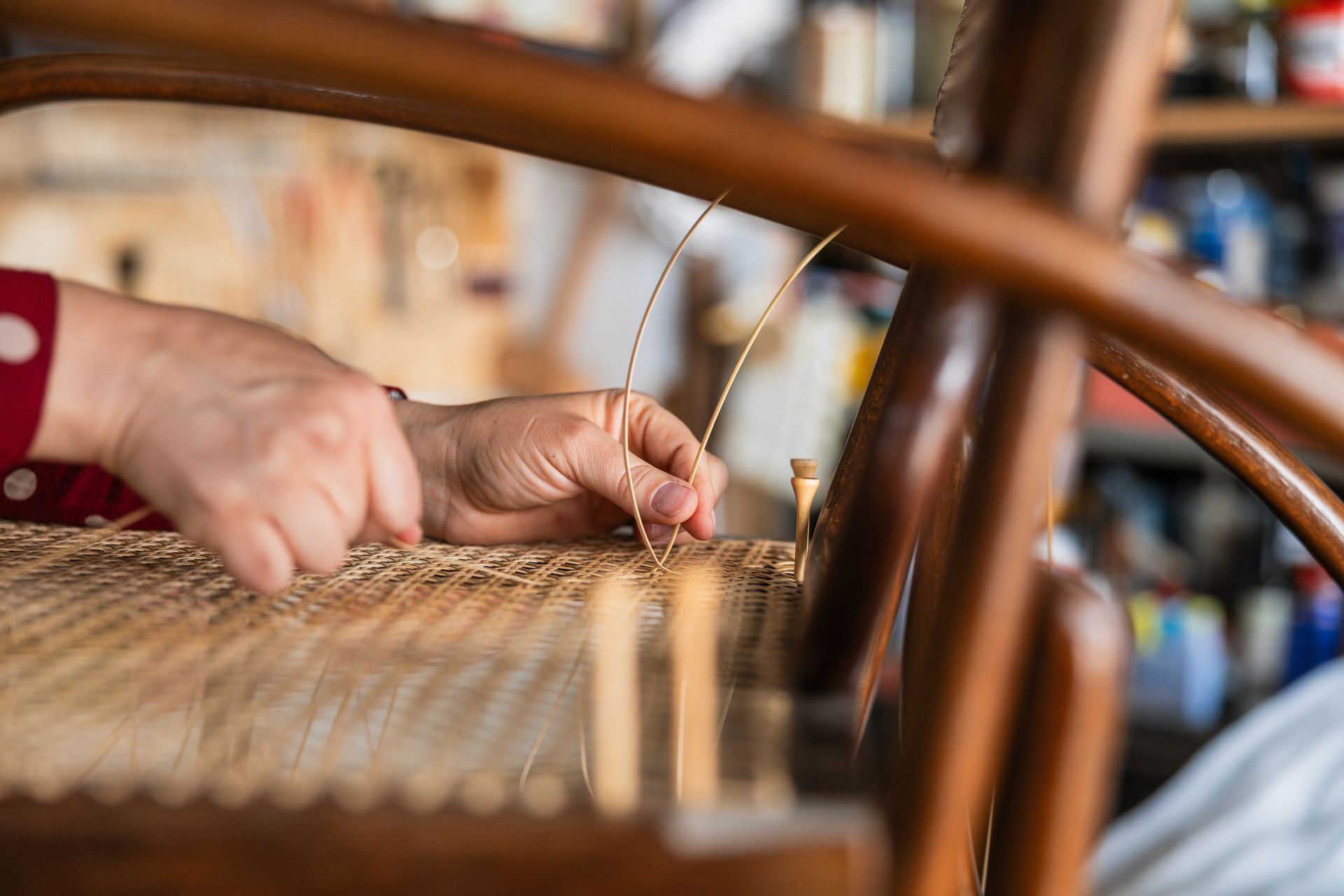 Close-up of hands weaving a rattan cane seat into a wooden chair frame in a workshop.
