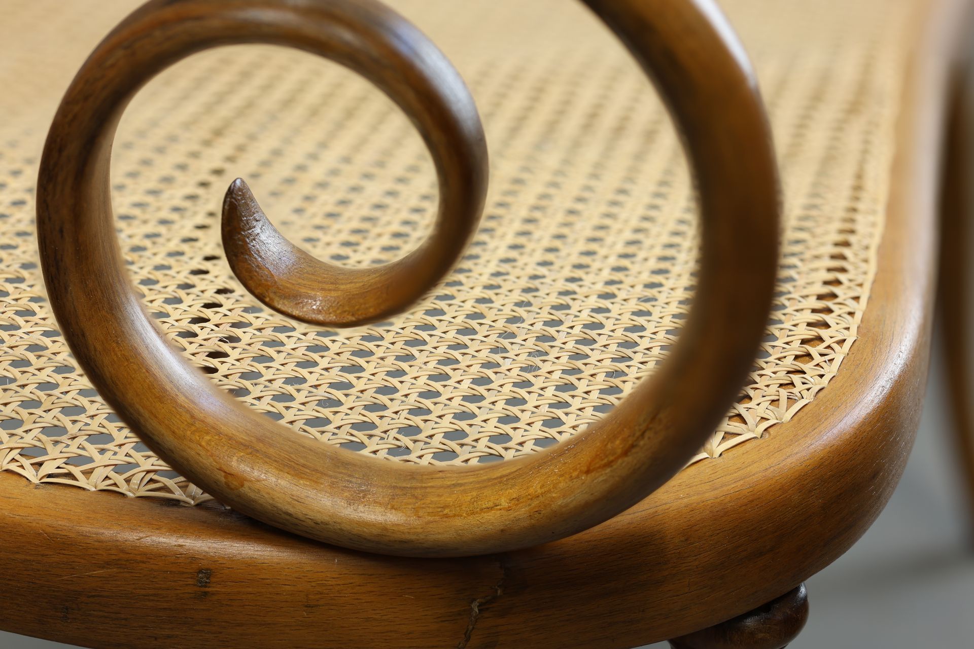 Close-up of a wooden chair armrest curled into a spiral, positioned above a woven cane seat.