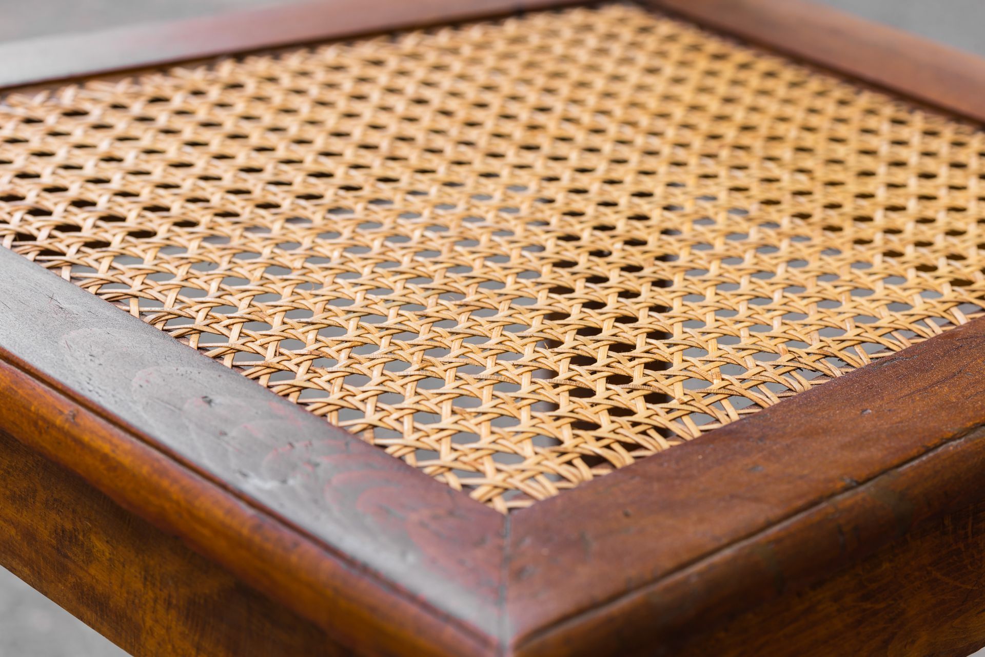 A close-up of a square wooden stool seat featuring a woven cane pattern in a neutral light brown color.