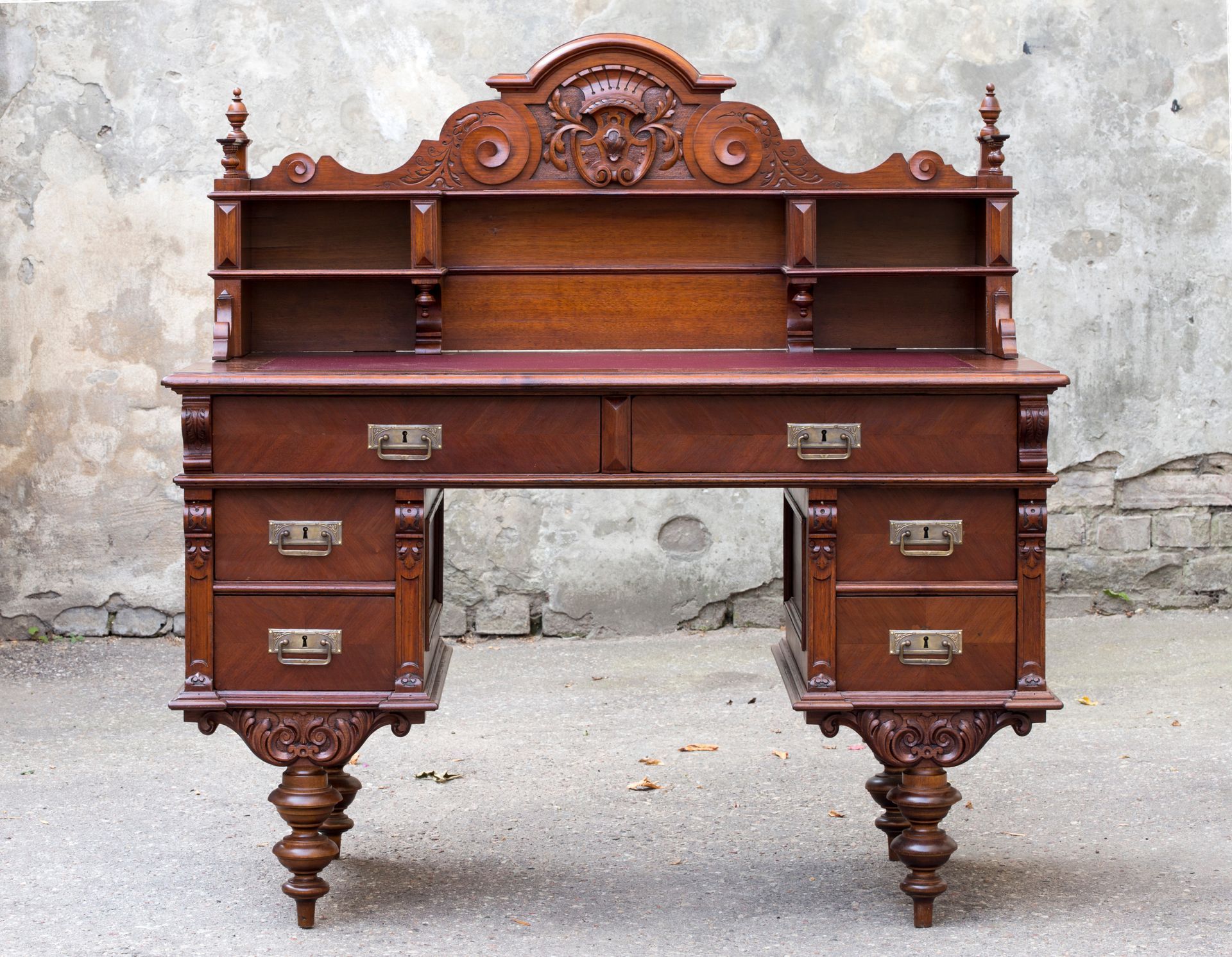 An ornate, dark-wood antique desk with a carved crest, shelving, two central drawers, and four side drawers on turned legs.