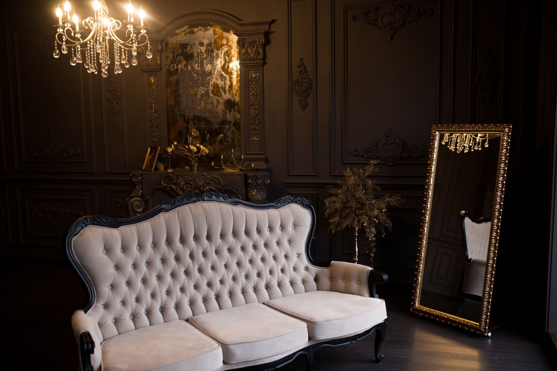 A light-colored tufted sofa sits in a dark, elegant room with a crystal chandelier and a tall gold-framed floor mirror.