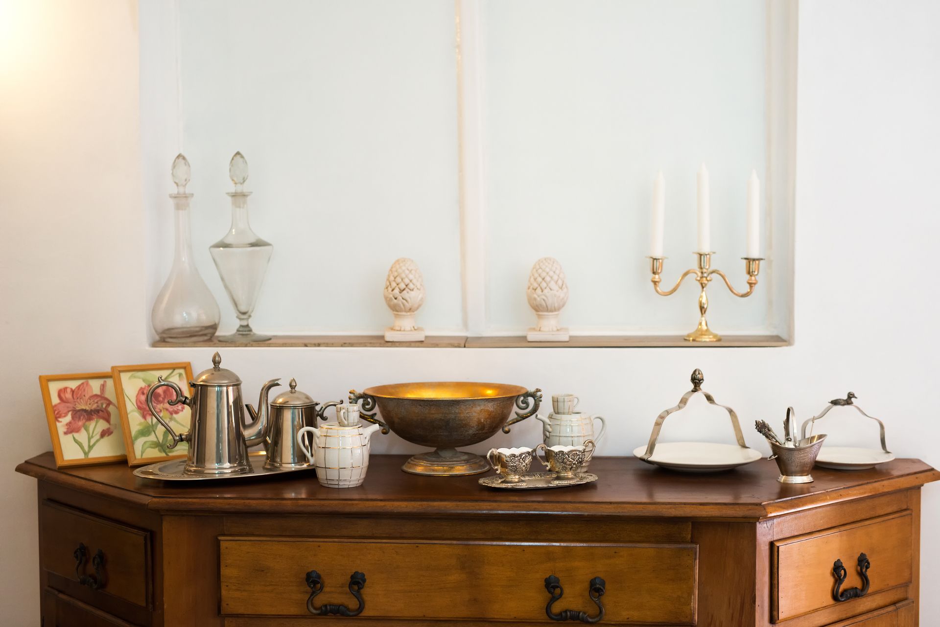 A wooden sideboard displays vintage silver teaware, floral prints, a gold bowl, candles, and glass decanters.