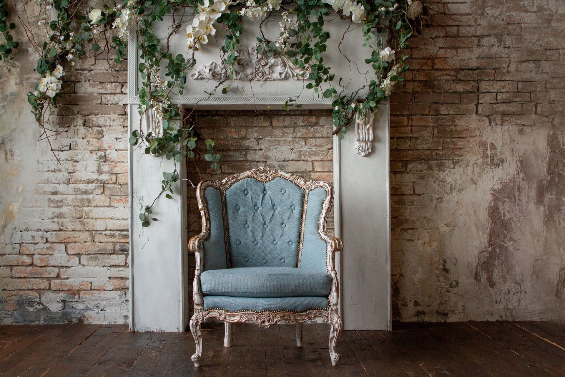 A light blue vintage armchair sits before a white fireplace mantel adorned with greenery against a weathered brick wall.