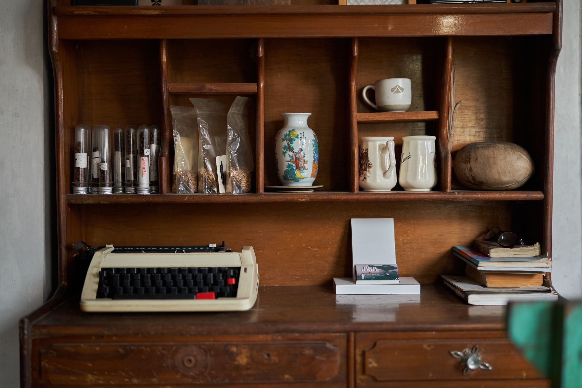An antique wooden hutch displaying a typewriter, small ceramic containers, a vase, mugs, and a stack of books.