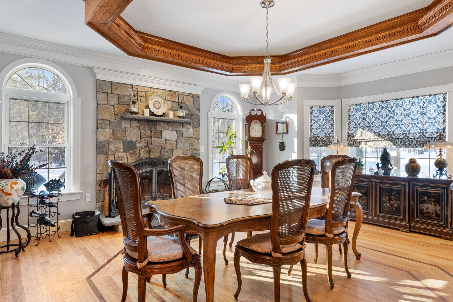A formal dining room features a wooden table with chairs, a stone fireplace, and a large chandelier under a tray ceiling.