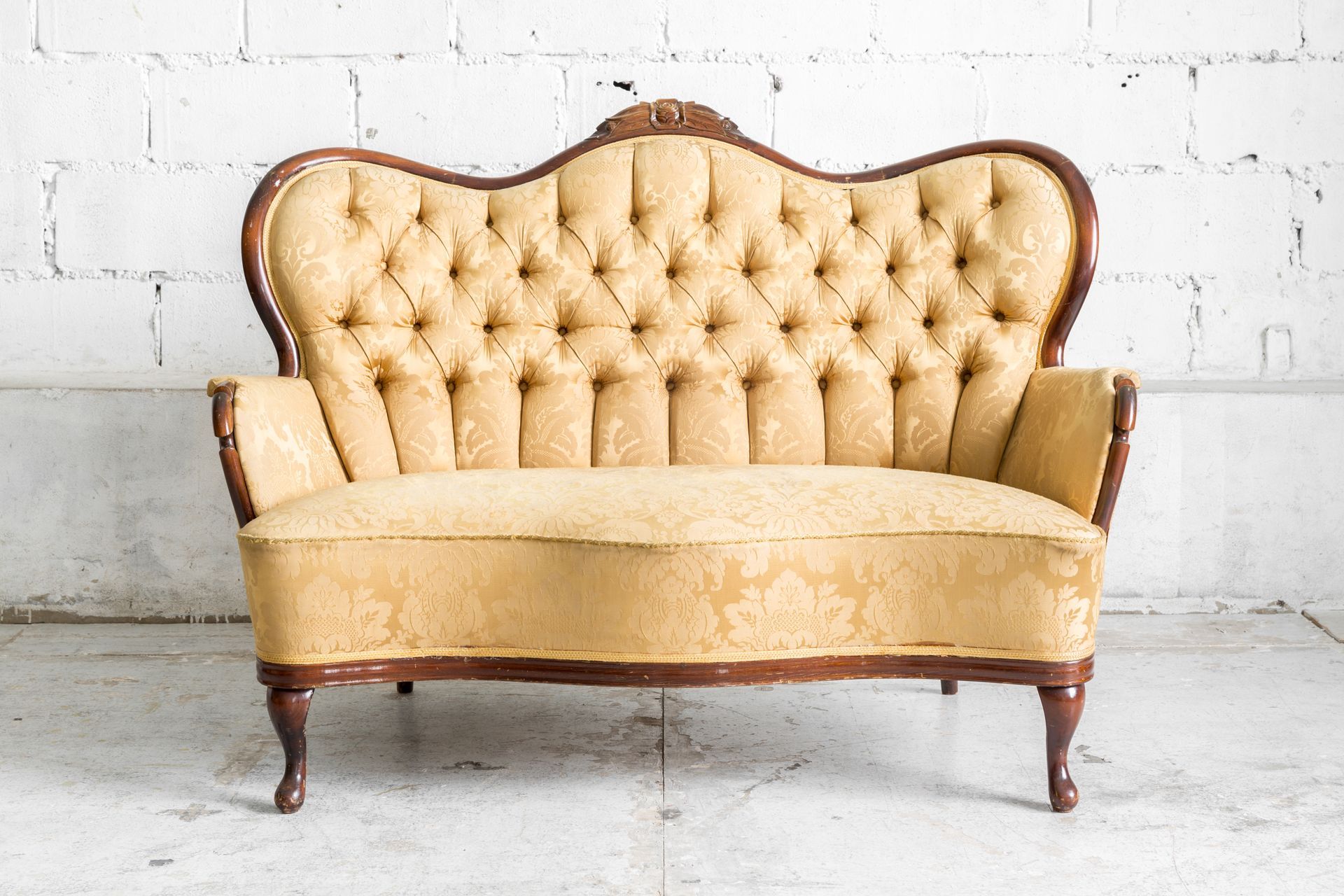 A vintage-style, tufted, pale yellow sofa with dark wood trim, set against a white brick wall.