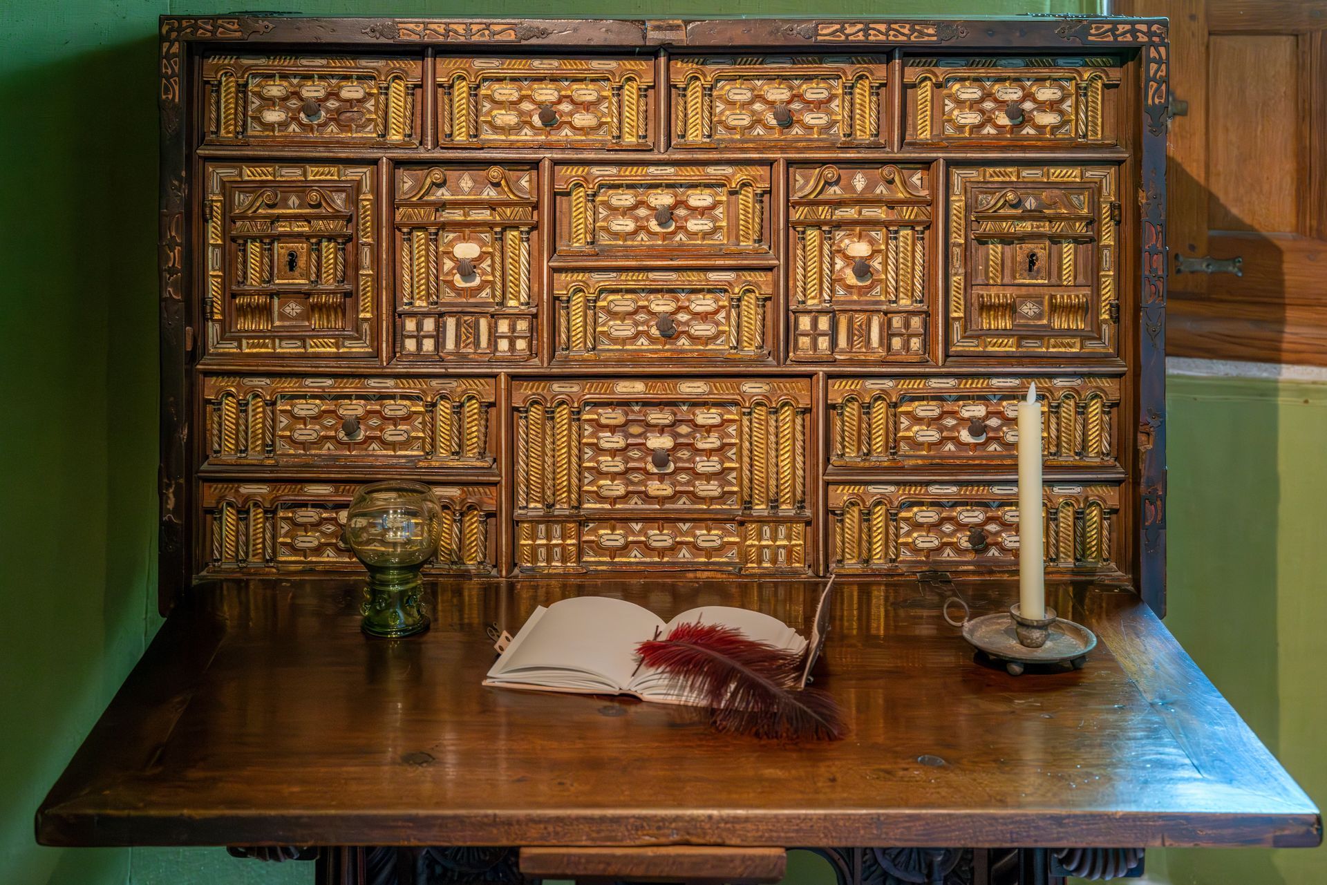 An antique wooden desk with ornate drawers, an open book, a red feather quill, and a candle on a green-walled background.