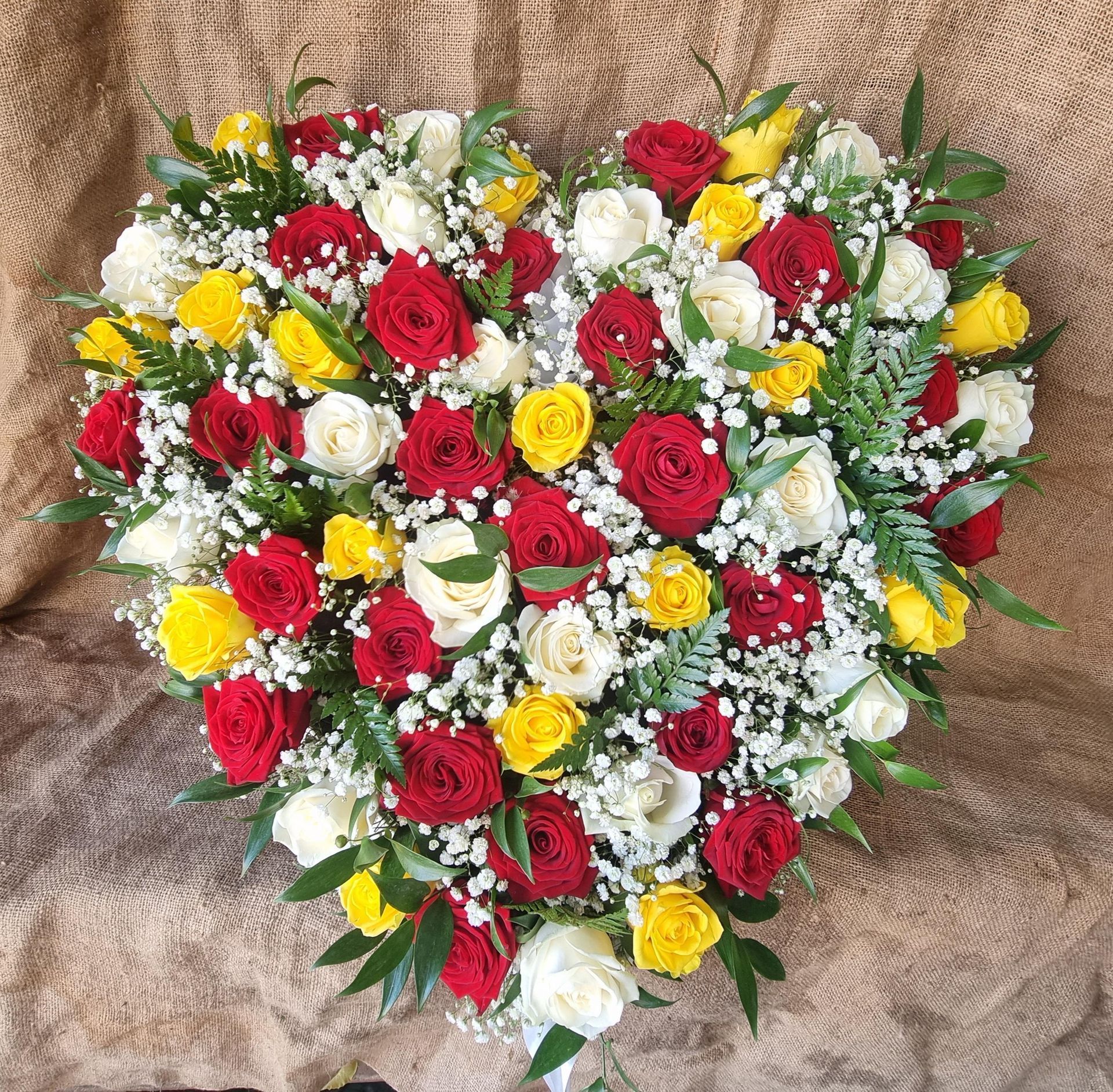 Heart-shaped floral arrangement with red, yellow, and white roses, surrounded by greenery, against a tan backdrop.