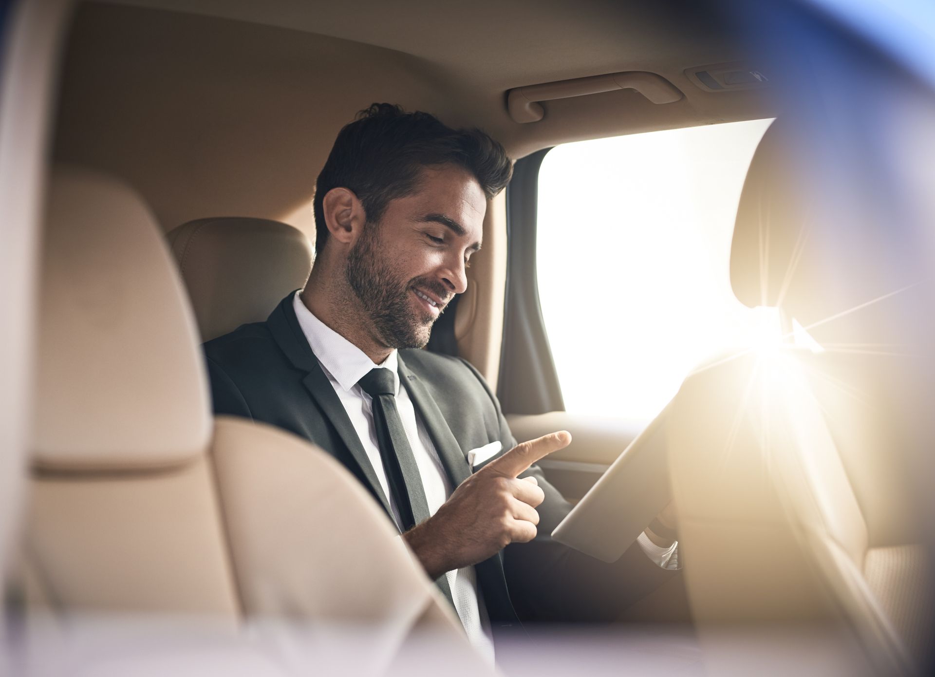 A man in a suit and tie is sitting in the back seat of a car using a tablet.
