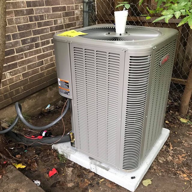 A gray air conditioner is sitting on a white block in front of a brick building