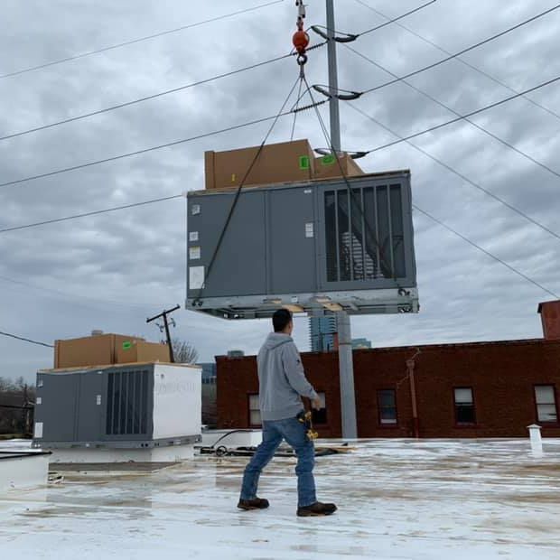 A man is walking on a snowy roof while a crane lifts a box with aircon on it