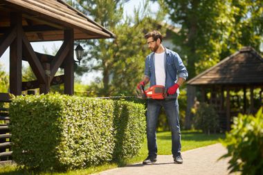 Man trimming hedges with a power tool in a garden, wearing gloves, near a gazebo.