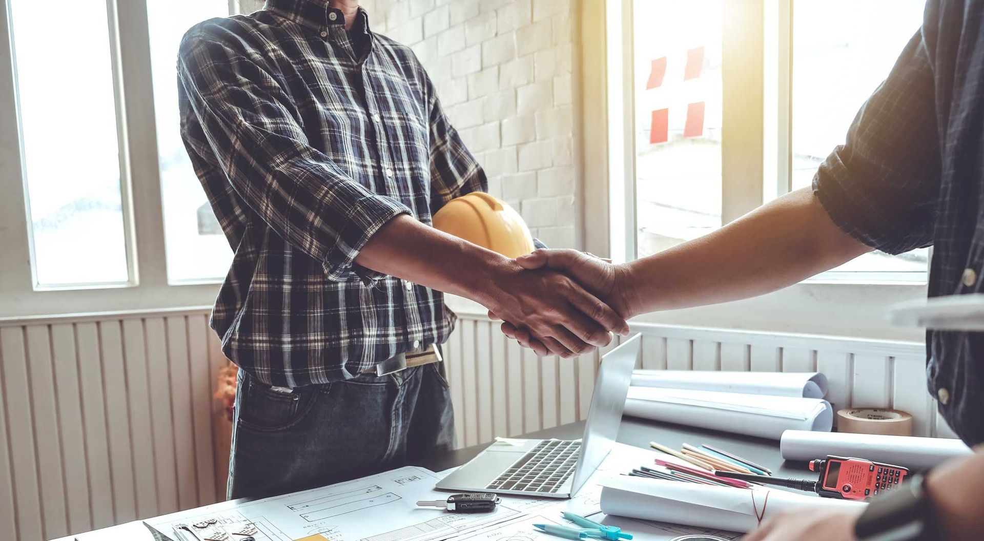 Two people shaking hands over a desk with construction plans and tools.