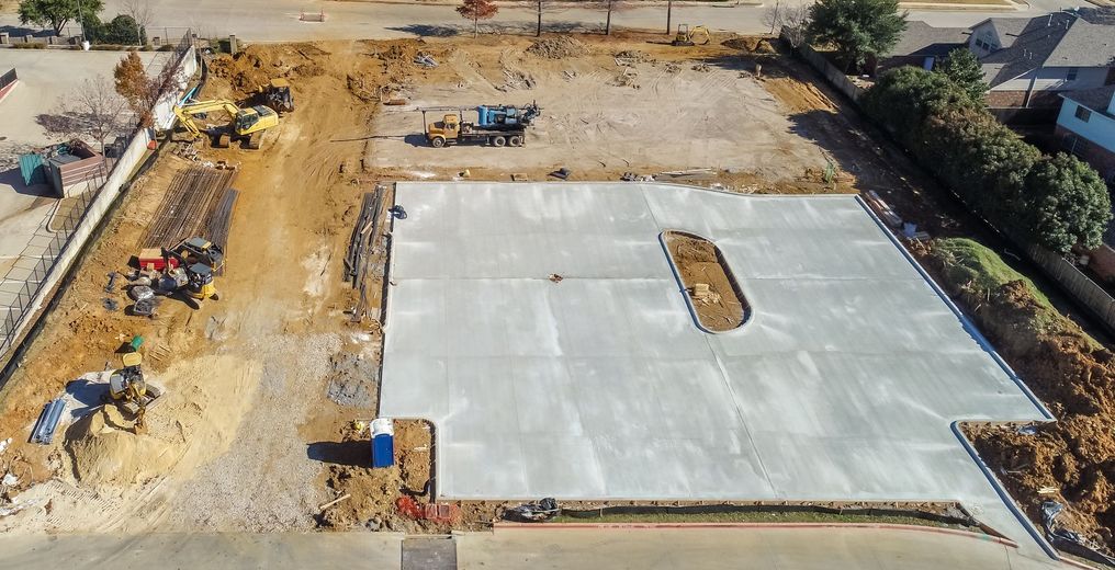 Aerial view of a construction site with concrete foundation, earth-moving equipment, and cleared land.