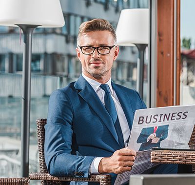 A professional wearing a suit and glasses sits at a table outdoors, looking forward while holding a business newspaper.