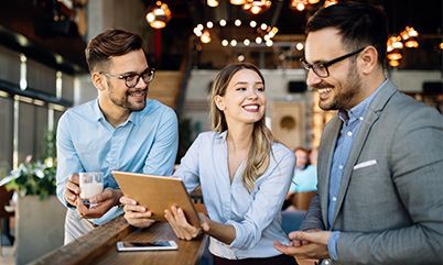 Three colleagues in business-casual attire smile and talk while gathered around a tablet at an indoor wooden counter.