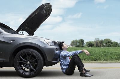 A person in business attire sits on the road, leaning against a grey car with an open hood, on a roadside near a field.