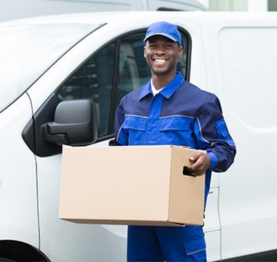 A smiling delivery worker in a blue uniform holds a cardboard box in front of a white delivery van.