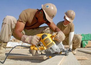 Two construction workers in tan uniforms and hard hats operate a yellow circular saw on a wooden board outdoors.